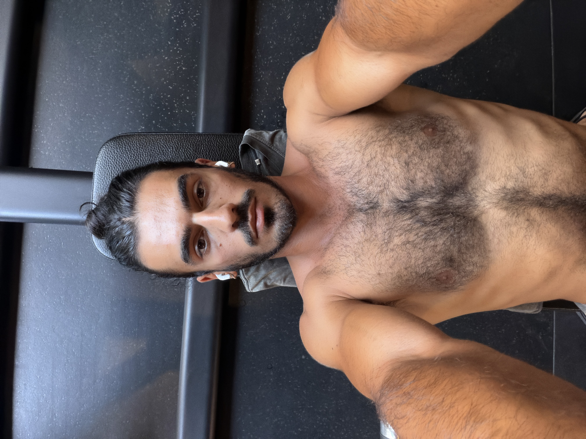 A shirtless man with a mustache and a hair bun lies on a black leather surface, possibly a weight bench. He appears to be taking a selfie, as his arms are extended toward the camera. He is wearing what appears to be wireless earbuds. The background includes a black wall or surface with a speckled pattern, possibly the floor or wall of a gym. There is no indication of the location being North Kuta, Indonesia.