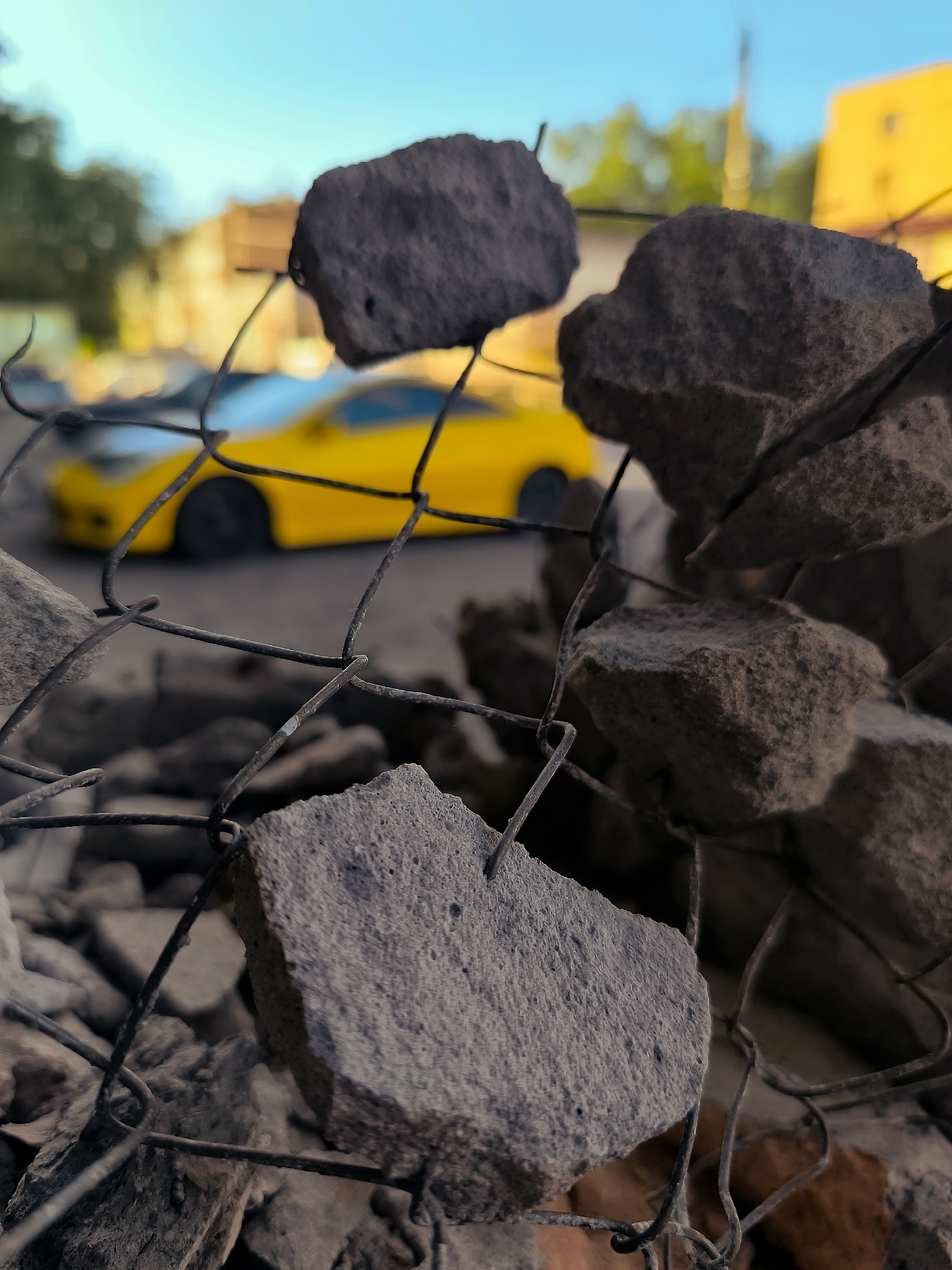 A low-angle, close-up shot captures a section of a wire fence with sharp, jagged pieces of concrete and rubble attached and in front of it. In the background, out of focus, a bright yellow sports car is parked on a paved surface. Beyond the car, blurred buildings and trees are visible under a clear blue sky. The scene appears to be an urban setting, possibly in Qarağandı city administration, Kazakhstan.