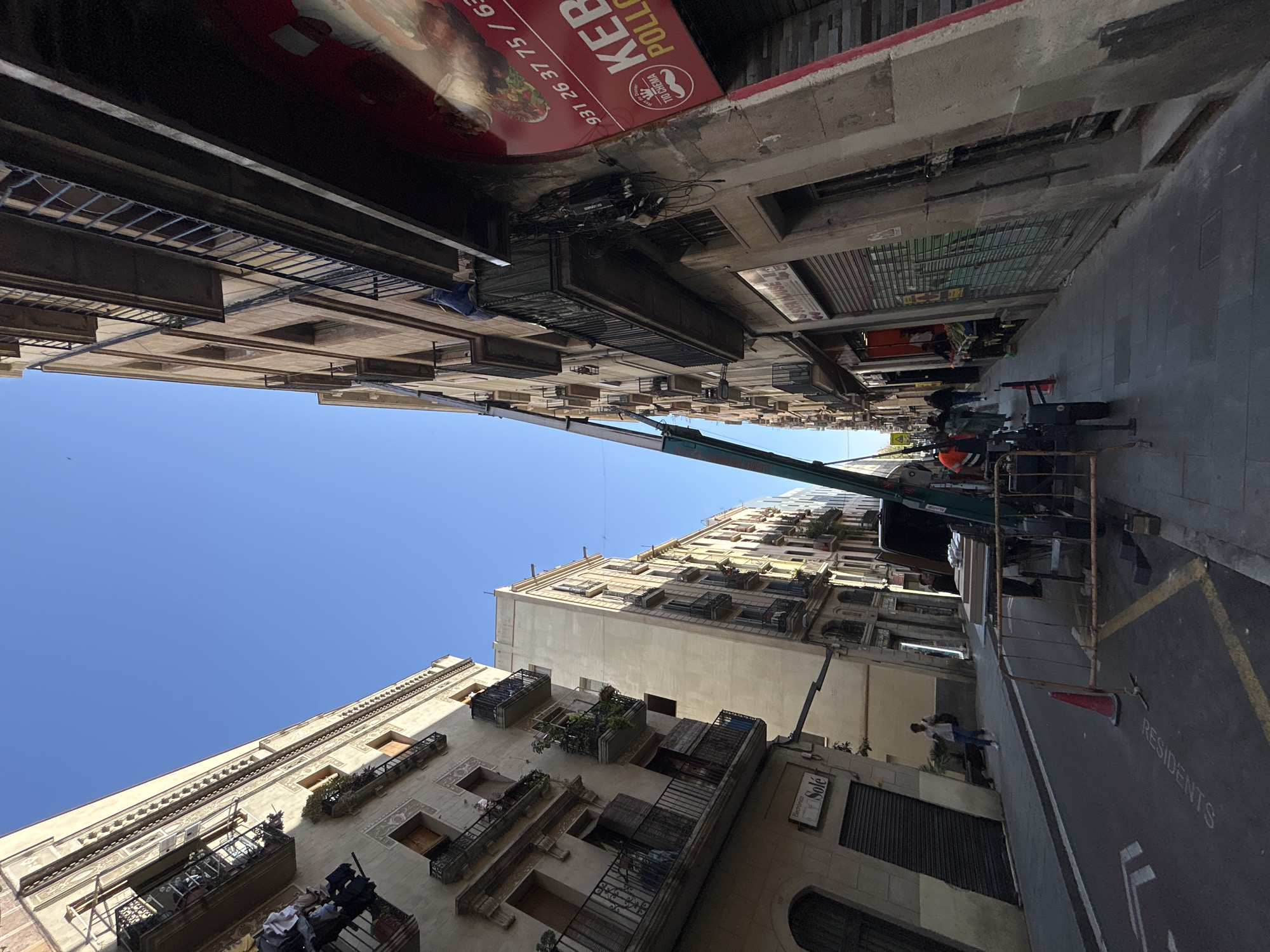 Captured at an unusual angle, the image reveals an urban scene in Barcelona, Spain. The perspective is tilted, with buildings lining both sides of a narrow street, their architecture characterized by multiple balconies and decorative elements. A construction crane is stationed in the street, its boom extended towards the upper stories of a building, suggesting ongoing work or maintenance. People are present, some appearing to be workers near the crane, and others walking along the sidewalk. The street is marked with a designated zone for "RESIDENTS." A "KEBAB" sign is visible on one side of the street, indicating a local business. The sky is clear and blue.
