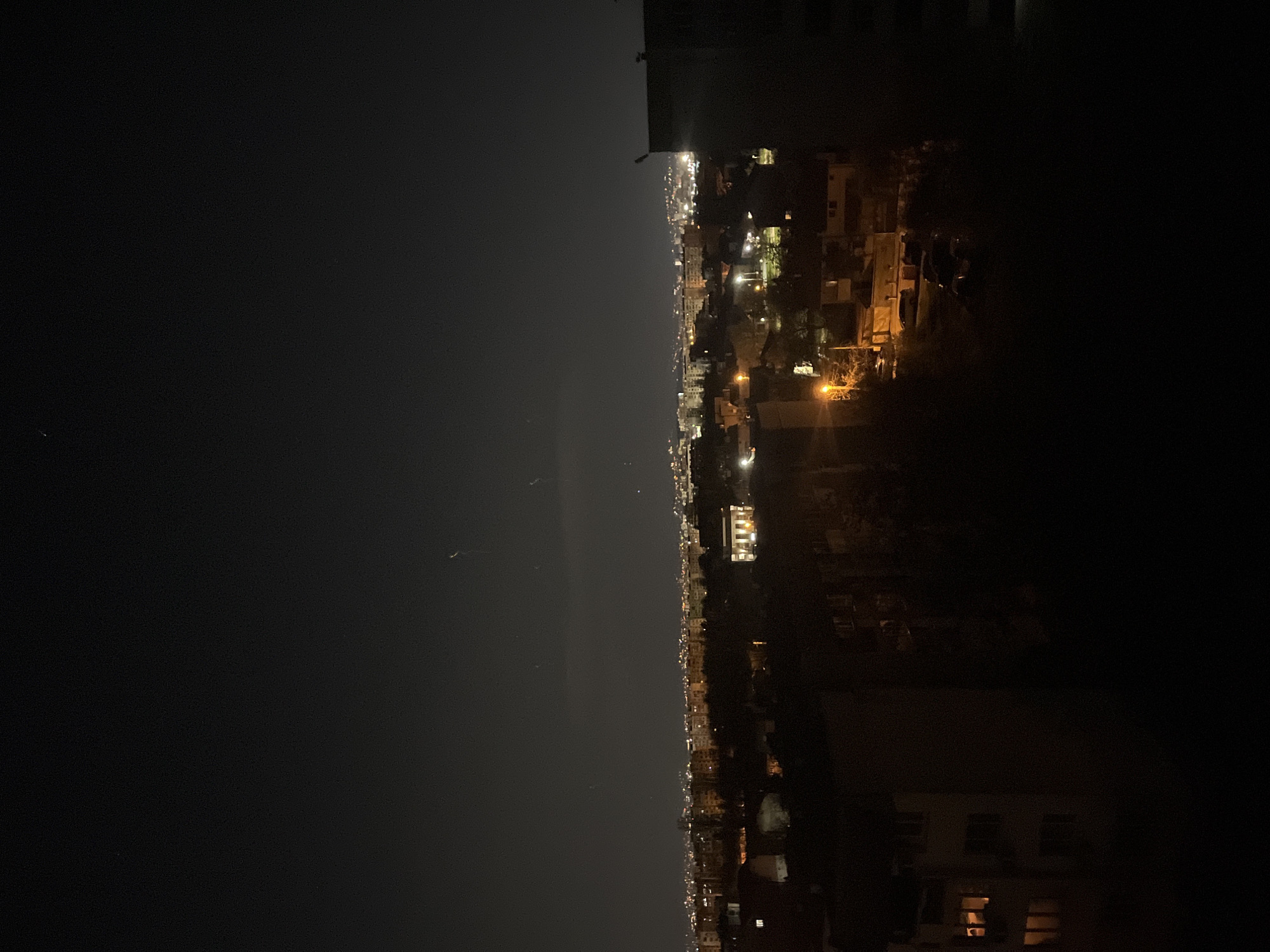 The image shows a nighttime cityscape in Sofia, Bulgaria. The viewpoint is elevated, likely from a high-rise building. The city lights extend horizontally across the central part of the image. Buildings and structures are visible on both the left and right edges, with details obscured by darkness. The left side of the image is mostly dark, suggesting the night sky.