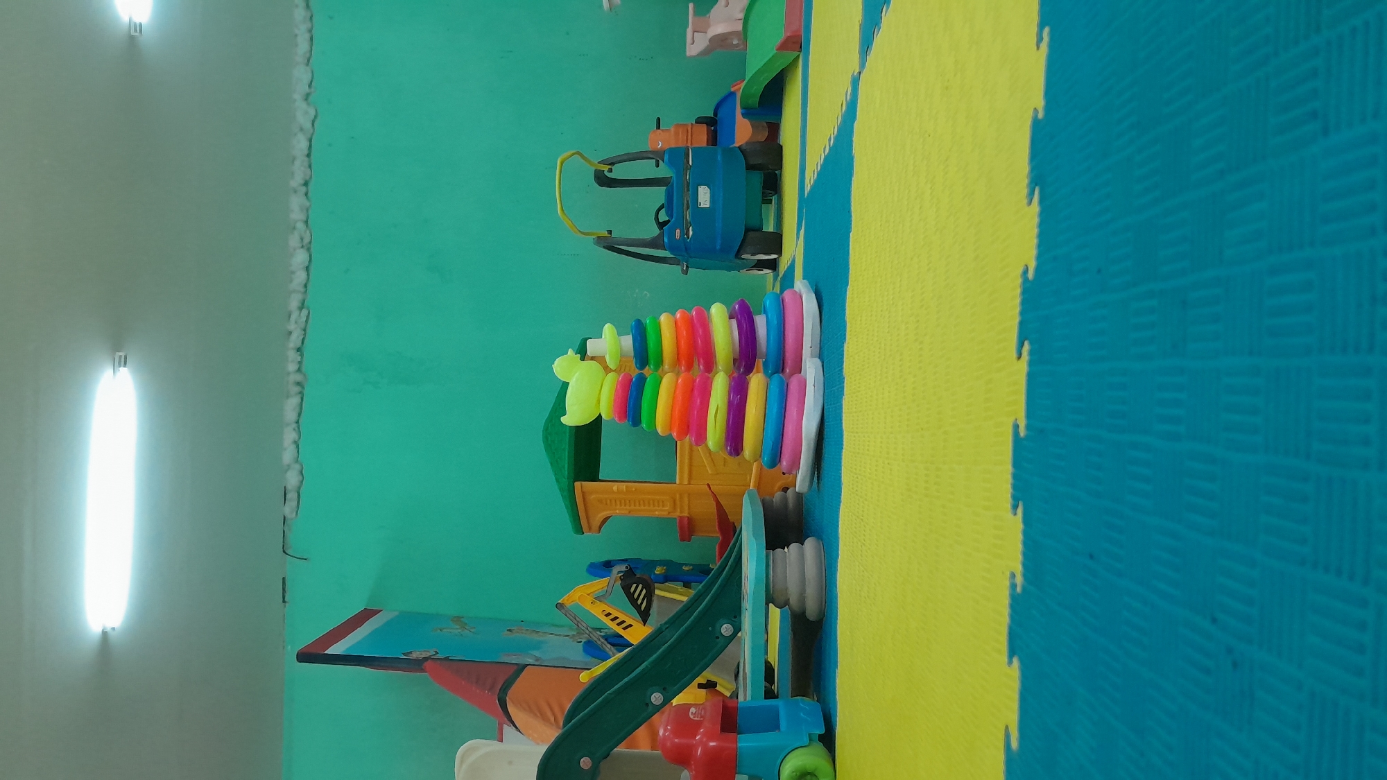 The image depicts an indoor children's play area with brightly colored toys and a turquoise-painted wall. In the foreground, a floor mat is composed of interlocking yellow and blue puzzle pieces. A small red and blue toy car is positioned on the left, next to a green slide supported by gray cylindrical bases. Behind the slide, a yellow structure resembling a playhouse with a green roof is visible. To the right of the playhouse, two stacks of colorful stacking rings are arranged, topped with a yellow duck figure. Further to the right, a blue ride-on car with a yellow handle is parked. In the background, a portion of a large, colorful play structure or mural is visible on the left side. Two fluorescent lights are mounted on the ceiling, illuminating the space. The environment appears to be a public or institutional setting, possibly a daycare or playroom in the city of Kasiman, Indonesia. There are no people present in the image.