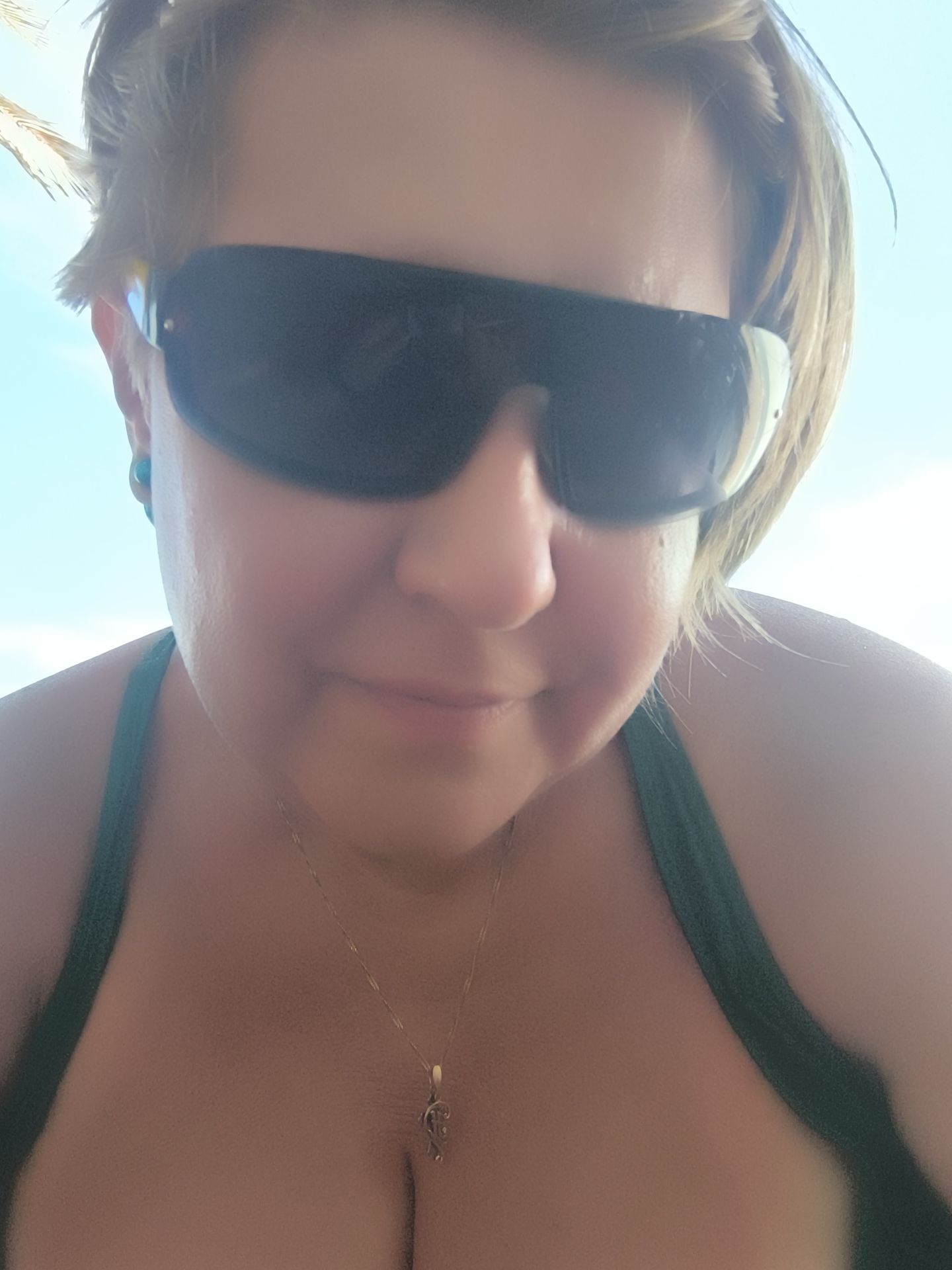 A woman is taking a selfie outdoors. She is wearing sunglasses and a green bikini top. A gold necklace with a small pendant is visible. The background is a bright blue sky. The lighting suggests it is daytime. The image appears to have been taken in Phu Thuy Ward, Vietnam.