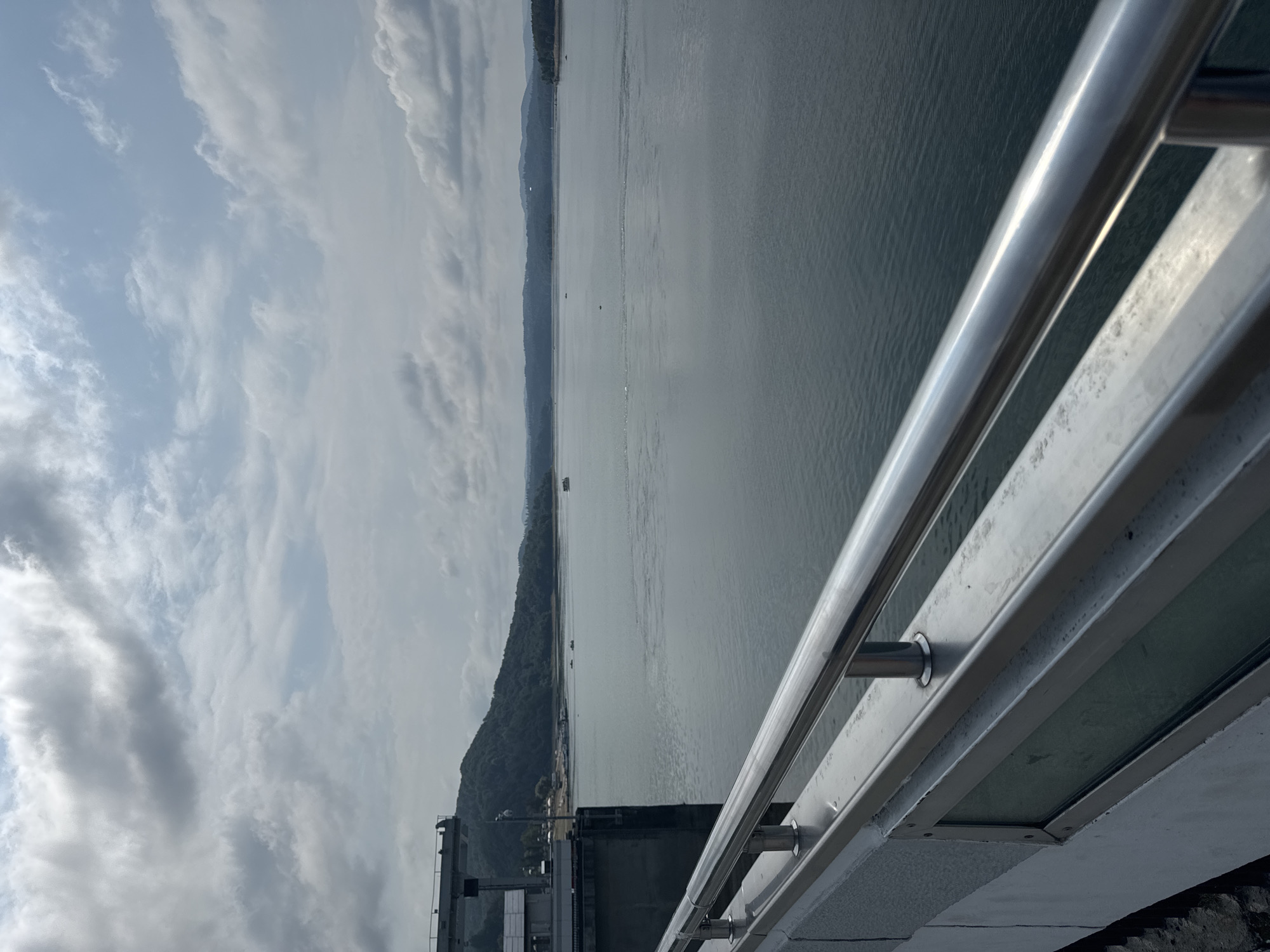A wide-angle, horizontal, eye-level shot captures a serene view of the Solina Lake from the Solina Dam in Solina, Poland. The foreground is dominated by the metallic railing of a walkway, constructed with polished stainless steel tubes and gray concrete supports. To the left, a portion of the dam's structure is visible, featuring industrial components and a building.
The vast expanse of the lake stretches into the distance, its surface reflecting the overcast sky. The water is calm, with subtle ripples indicating slight movement. Several small boats are scattered across the lake, some appearing to be fishing or leisure craft. In the background, a line of verdant hills meets the horizon, covered in dense forestation. The sky above is filled with a mix of gray and white clouds, with occasional patches of lighter blue. The overall atmosphere is one of tranquility and natural beauty, accentuated by the imposing presence of the dam.