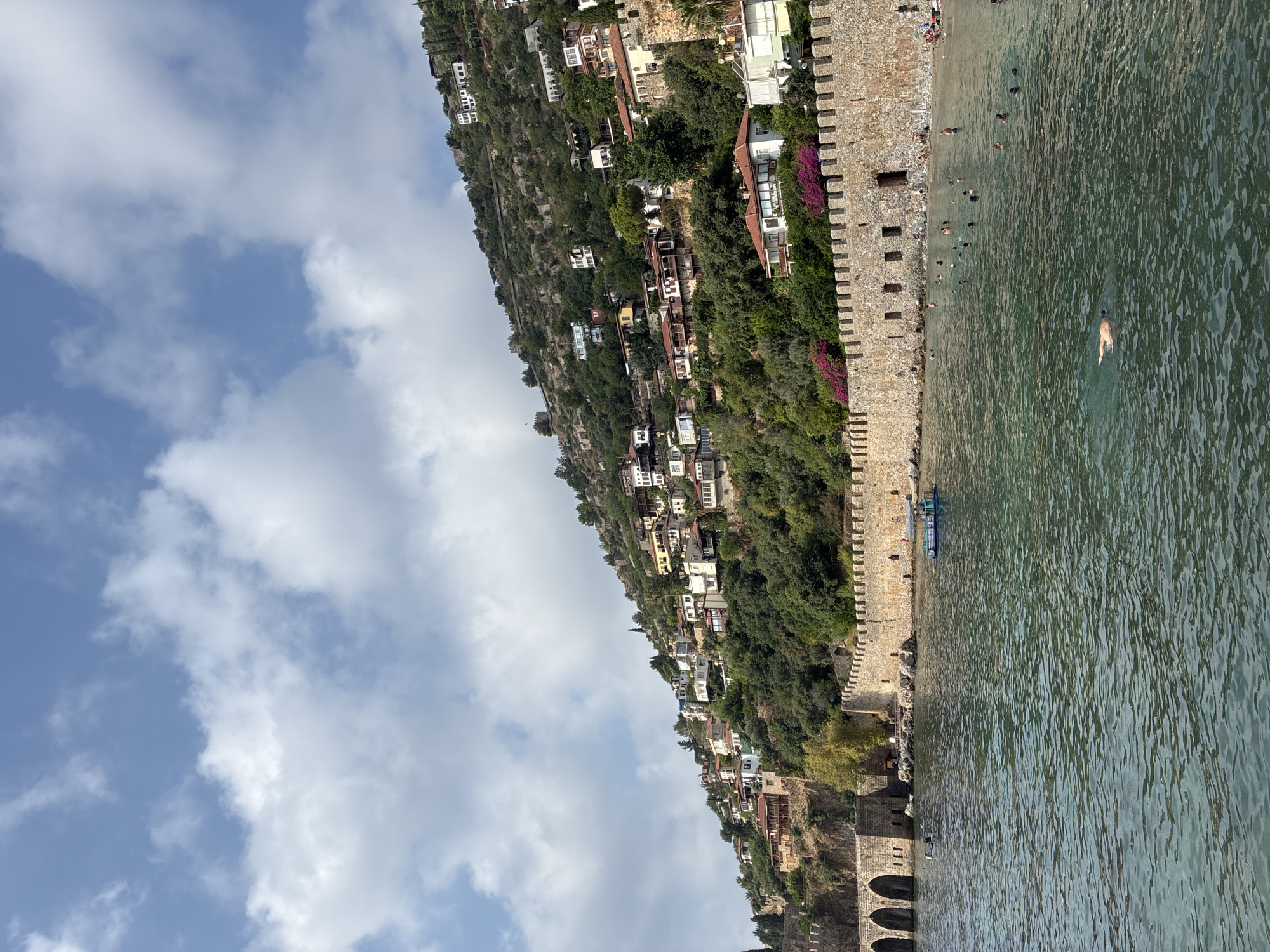 This is a photograph taken from the sea, looking towards a coastline in Alanya, Turkey. The scene depicts a hillside densely populated with traditional white and terracotta-roofed houses. A substantial stone wall, likely part of a historical fortification, runs along the base of the hill, separating it from the sea. The wall has crenellations and arched openings.
The sea in the foreground is a deep green, with visible ripples and reflections. Several people are visible in the water, swimming or floating. Some are close to the shore, while one individual is swimming further out. A small boat with a blue canopy is also in the water near the shore.
The beach area behind the stone wall appears to have several sunbathers and people walking. The hillside is covered in lush green vegetation, with trees and shrubs interspersed between the houses. The sky above is a bright blue with scattered white, fluffy clouds. The overall impression is a vibrant coastal scene with historical elements.