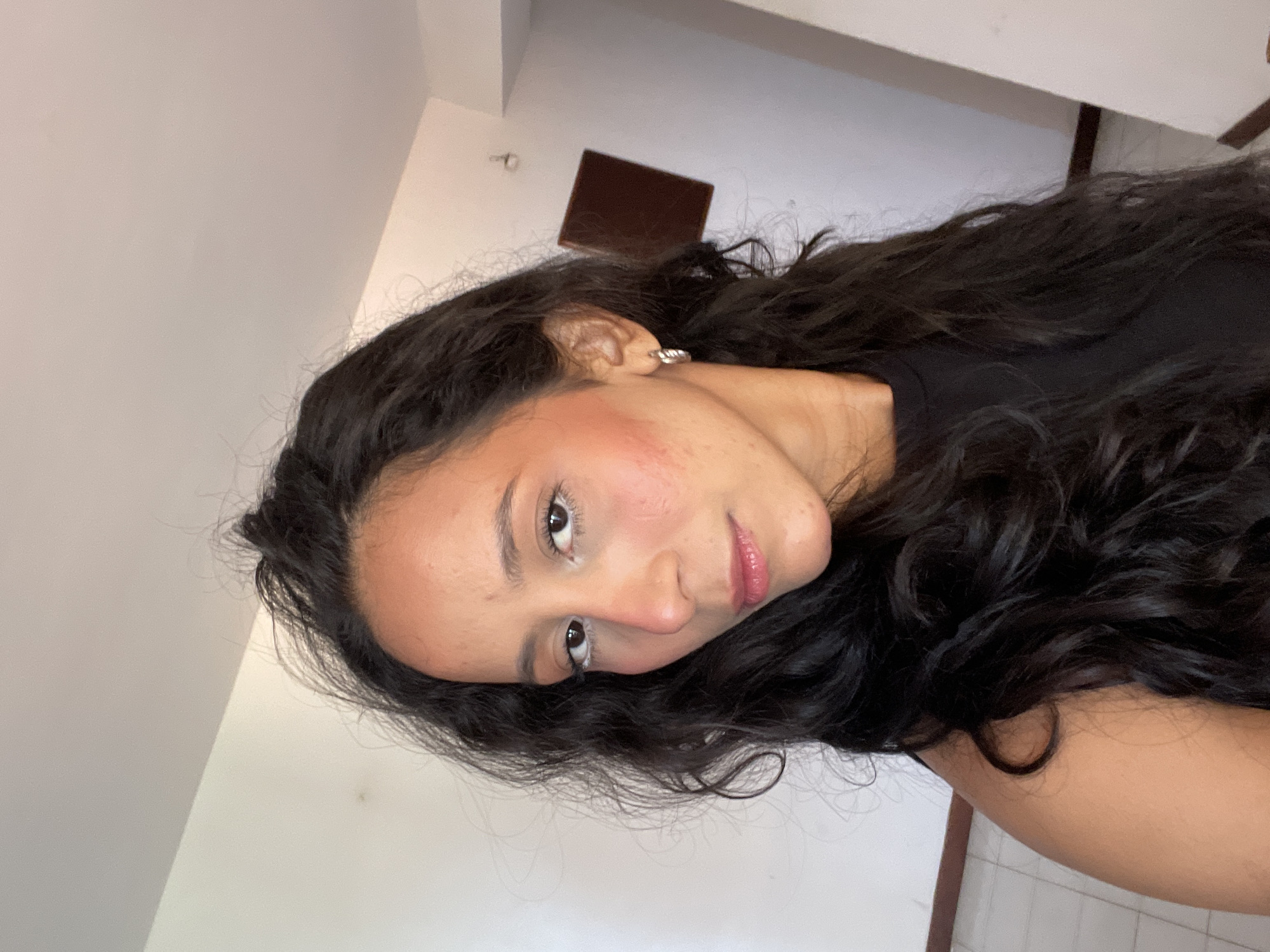 The image depicts a medium shot of a woman with long, dark, curly hair, taken from a slightly low angle. She is looking towards the camera with a neutral expression, her head tilted slightly to her left. Her skin appears to have some redness on her cheeks, possibly from blush or irritation. She is wearing minimal makeup, with a touch of white eyeliner on her lower waterline and lip gloss. A small silver hoop earring is visible in her left ear. She is wearing a black top. The background appears to be an indoor setting with white walls and a tiled floor. A brown square object is mounted on the wall in the distance, and a hook or nail is also visible on the wall. The lighting suggests an indoor environment, possibly during the daytime. The overall scene is simple and candid. Without specific landmarks or contextual cues within the image, it is impossible to definitively place the location in Porlamar, Venezuela.
