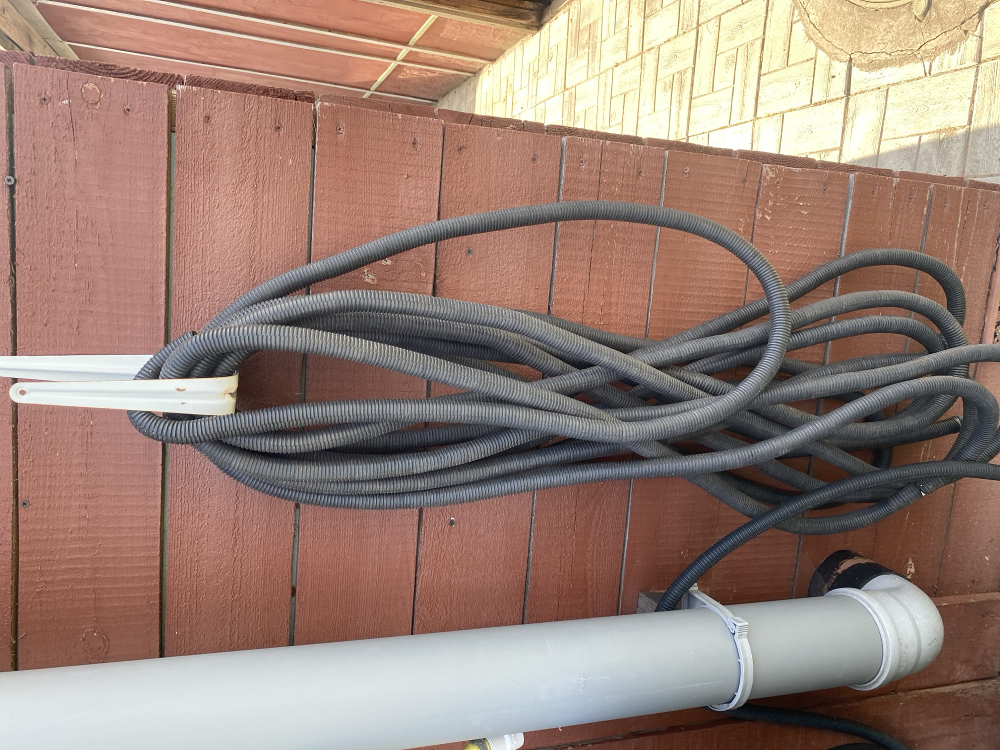 A coil of black corrugated tubing hangs from a white hook attached to a dark red wooden wall. To the left of the tubing is a light grey PVC pipe, which connects to a white elbow joint at the bottom, then transitions to black tubing that disappears behind the coiled black tubing. The wall is constructed of horizontal wooden planks. To the right of the wall, a path made of light-colored rectangular pavers is visible, leading into the distance where a wooden fence can be seen. The scene appears to be outdoors, with sunlight casting shadows on the pavers. The location is the city of Krupki, Belarus.