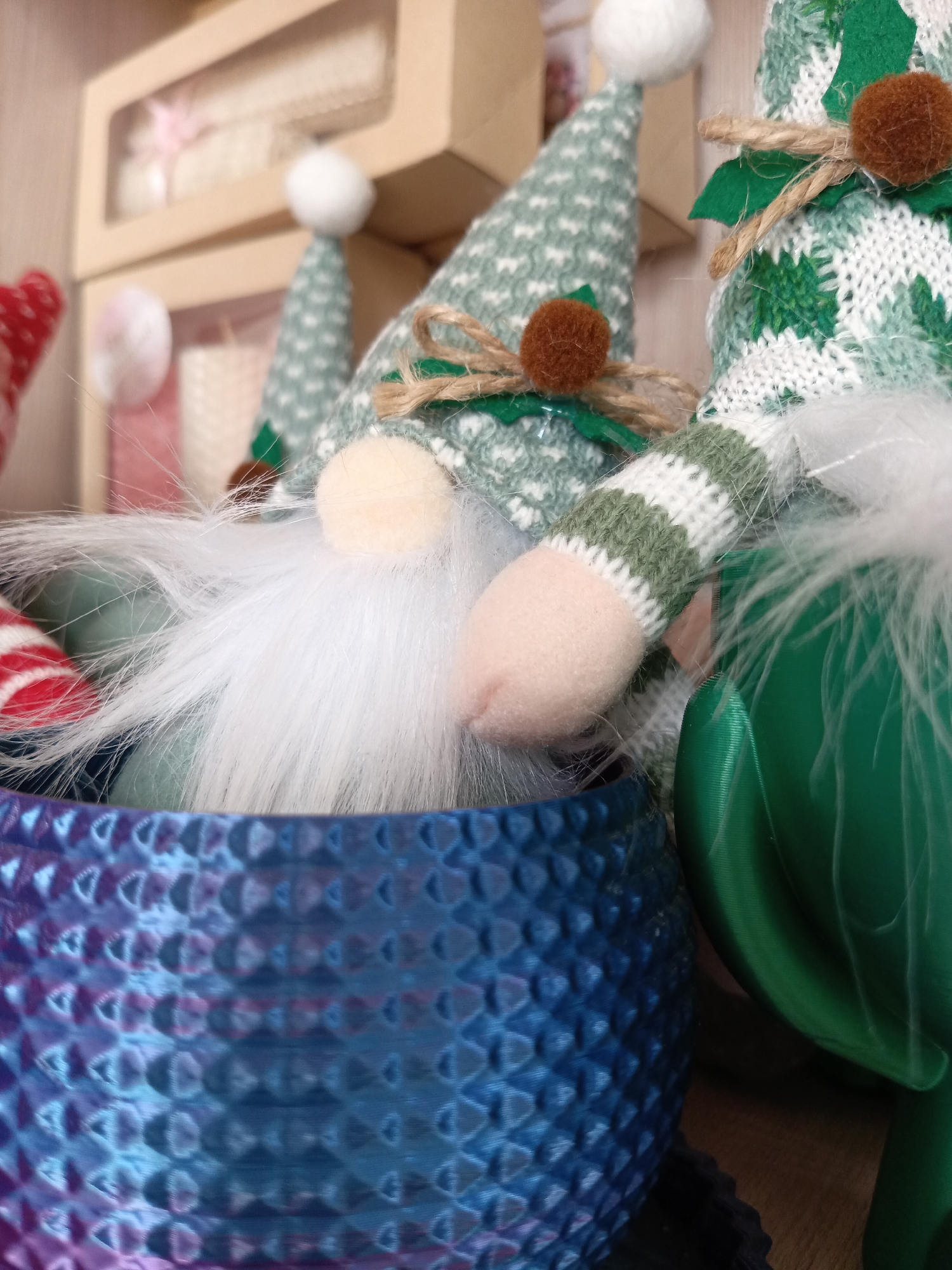 The image shows a close-up of several gnome figurines nestled inside a blue, textured pot. The gnomes have various green and white patterned hats, white beards, and pink noses. Some gnomes have brown pom-poms and green leaf details on their hats. The blue pot has a repeating circular pattern with a slight purple gradient at the bottom. The background includes blurred boxes, possibly containing more gnomes, and a wooden surface.