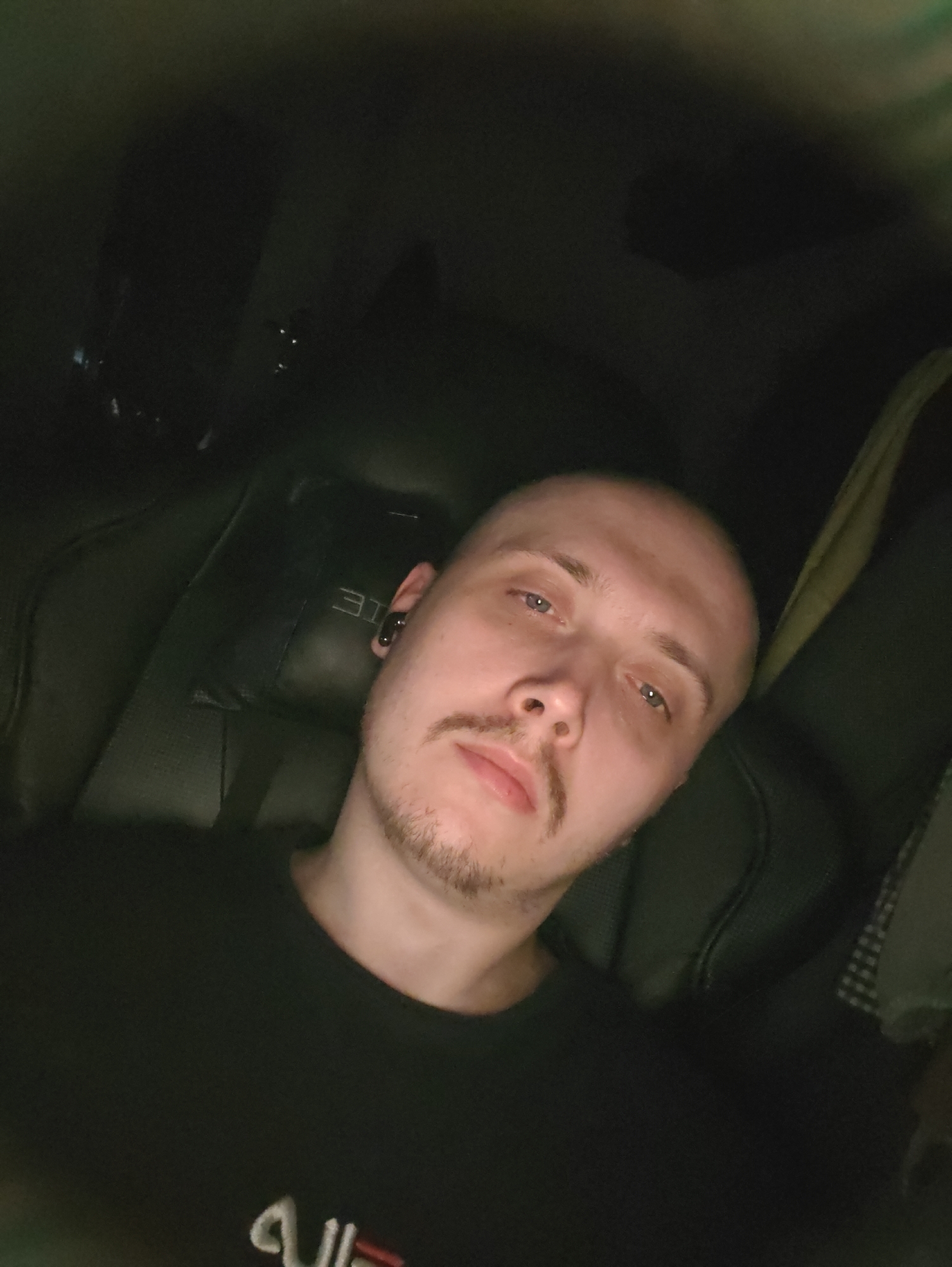 A close-up, slightly blurry, overhead shot captures a man from the chest up, seated in a dark interior, likely a vehicle. The man has a shaved head, light blue eyes, a small mustache, and a light goatee. He is wearing a black t-shirt with a white and red graphic on the left chest. He is also wearing a black wireless earbud in his right ear. The background is dark and indistinct, with a textured headrest visible behind him, appearing to have embossed lettering. The lighting is dim and appears to be coming from the front and slightly above the subject.