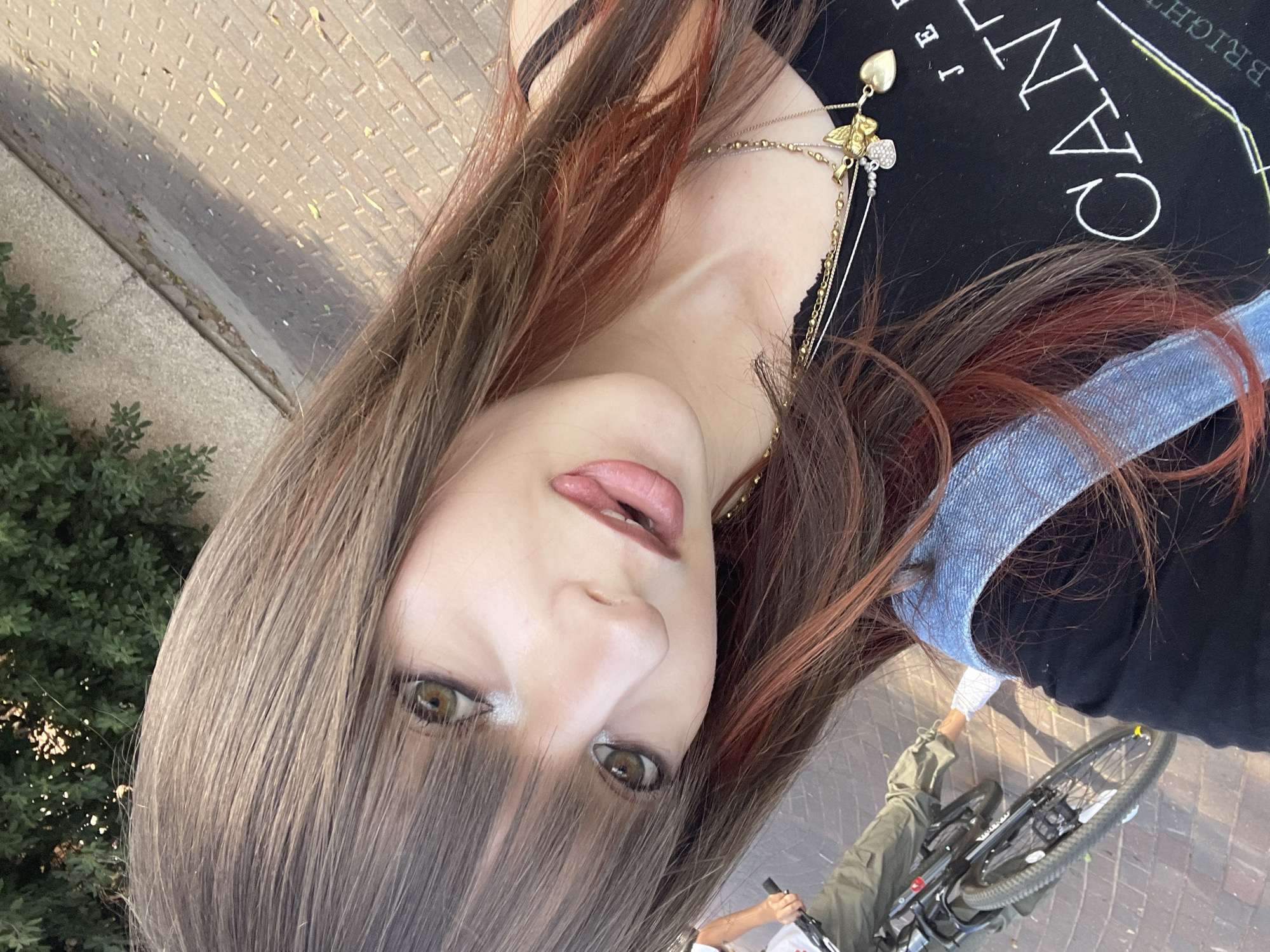 A close-up, low-angle selfie of a young woman with long, brown and auburn-streaked hair, bangs, and light makeup, looking at the camera with her mouth slightly open. She is wearing a black tank top with a graphic print and a denim vest over it, along with layered gold necklaces. In the background, out of focus, a person is riding a bicycle on a paved walkway next to a patch of green foliage. The scene appears to be outdoors on a sunny day.