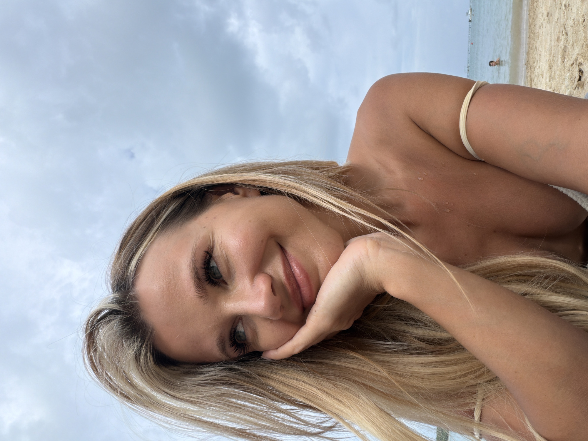 A young woman with blonde hair is photographed from the chest up while on a beach. She is resting her chin on her hand, looking off to the side with a slight smile. She is wearing a bikini top. In the background, the ocean is visible with a few boats and a person swimming. The sky is cloudy. The location is the city of Wichit, Thailand.