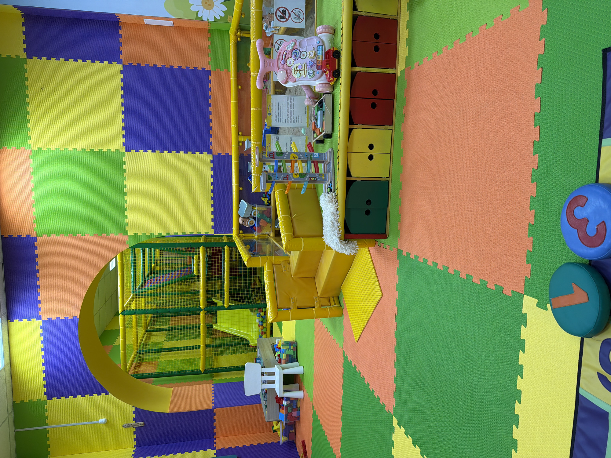 The image depicts an indoor children's play area. The floor is covered with interlocking foam mats in alternating colors of green, yellow, orange, and blue. On the right side of the image, large foam number shapes are visible: "1" and "3".
Various play structures are present, including a multi-level climbing frame enclosed with netting and featuring yellow support beams. A yellow platform with a small staircase leads to the climbing frame.
Various toys are present, including a pink walker toy and a wooden toy car ramp. Next to this is a storage unit with four levels of drawers in red, yellow, and green.
A small white child's table and chairs are positioned near the climbing frame, with blocks scattered around.
The image does not include any people. The location is not definitively identified, but the prompt suggests it is in Kamyshin, Russia.