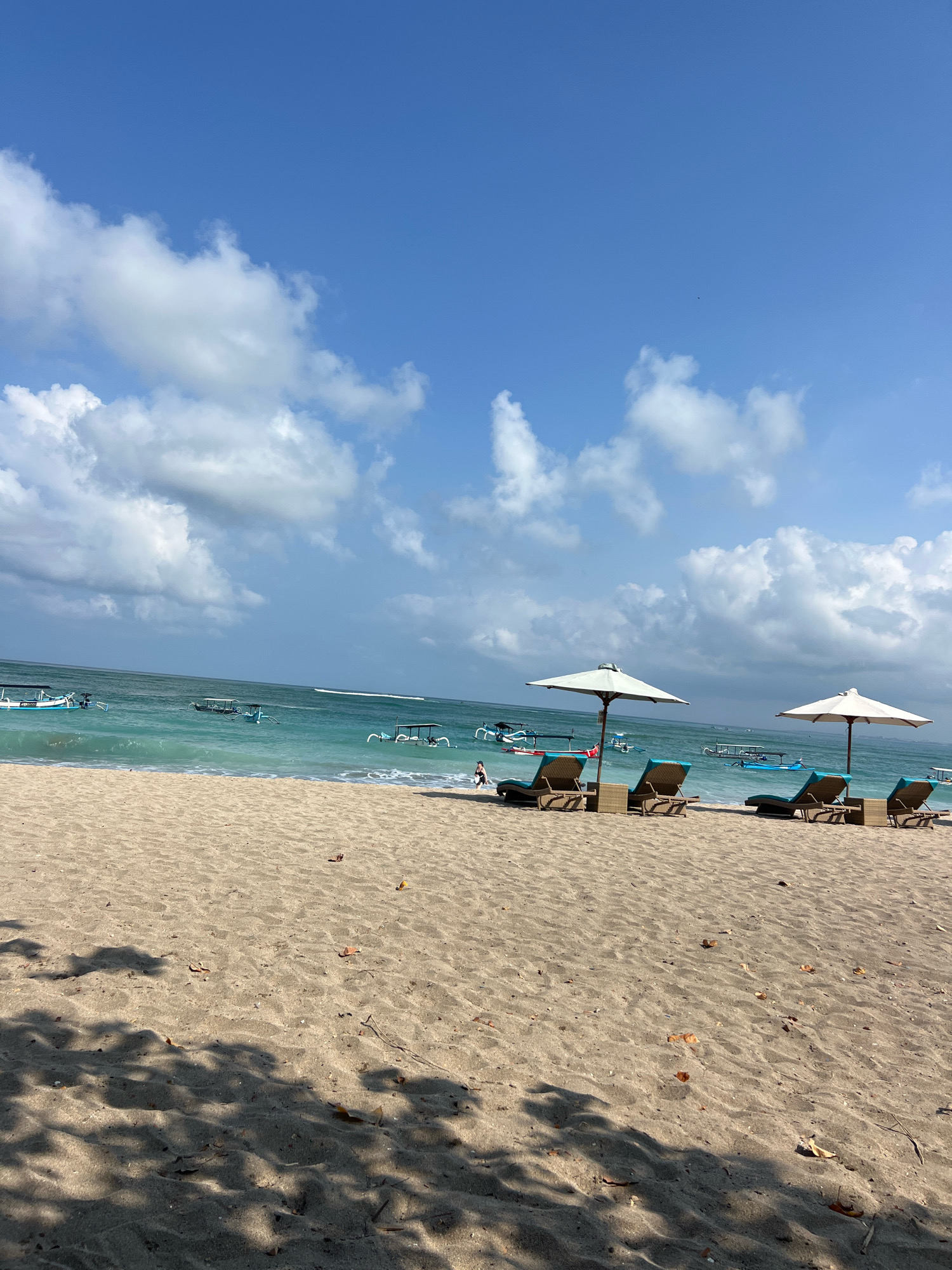 The image captures a serene beach scene in Kuta, Indonesia. The foreground is dominated by golden sand, marked with scattered dry leaves and shaded by dappled shadows from unseen trees. In the mid-ground, two pairs of lounge chairs with blue cushions are arranged under white umbrellas, facing the ocean. Beyond the chairs, the turquoise water of the sea stretches towards the horizon, dotted with several traditional outrigger boats. The sky above is a vibrant blue, adorned with scattered white cumulus clouds, creating a picturesque tropical setting. A lone figure is visible on the beach near the water's edge.