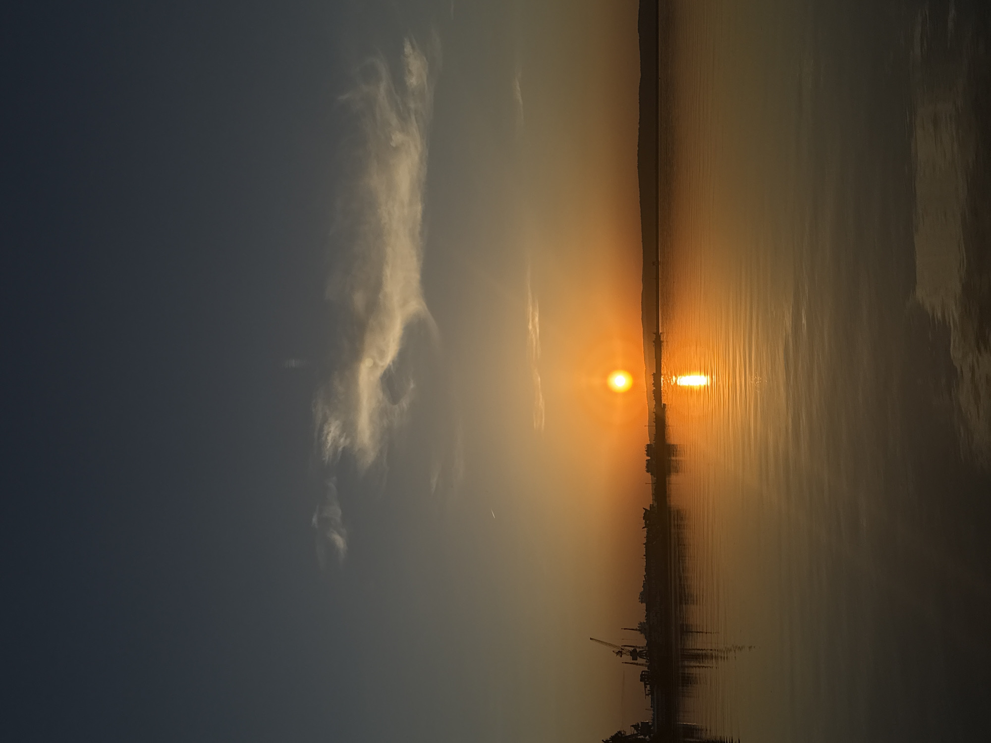 The image depicts a sunset over a body of water, likely a river or a bay, in the city of Chaikovsky, Russia. The sun is positioned low on the horizon, creating a bright reflection on the water's surface. The sky transitions from a dark blue on the left side to an orange-yellow hue near the horizon. Clouds are visible on the left side of the image. The coastline in the distance shows a silhouette of a city skyline with cranes, buildings, and industrial structures. The foreground features the dark surface of the water, with reflections of the sky and city.