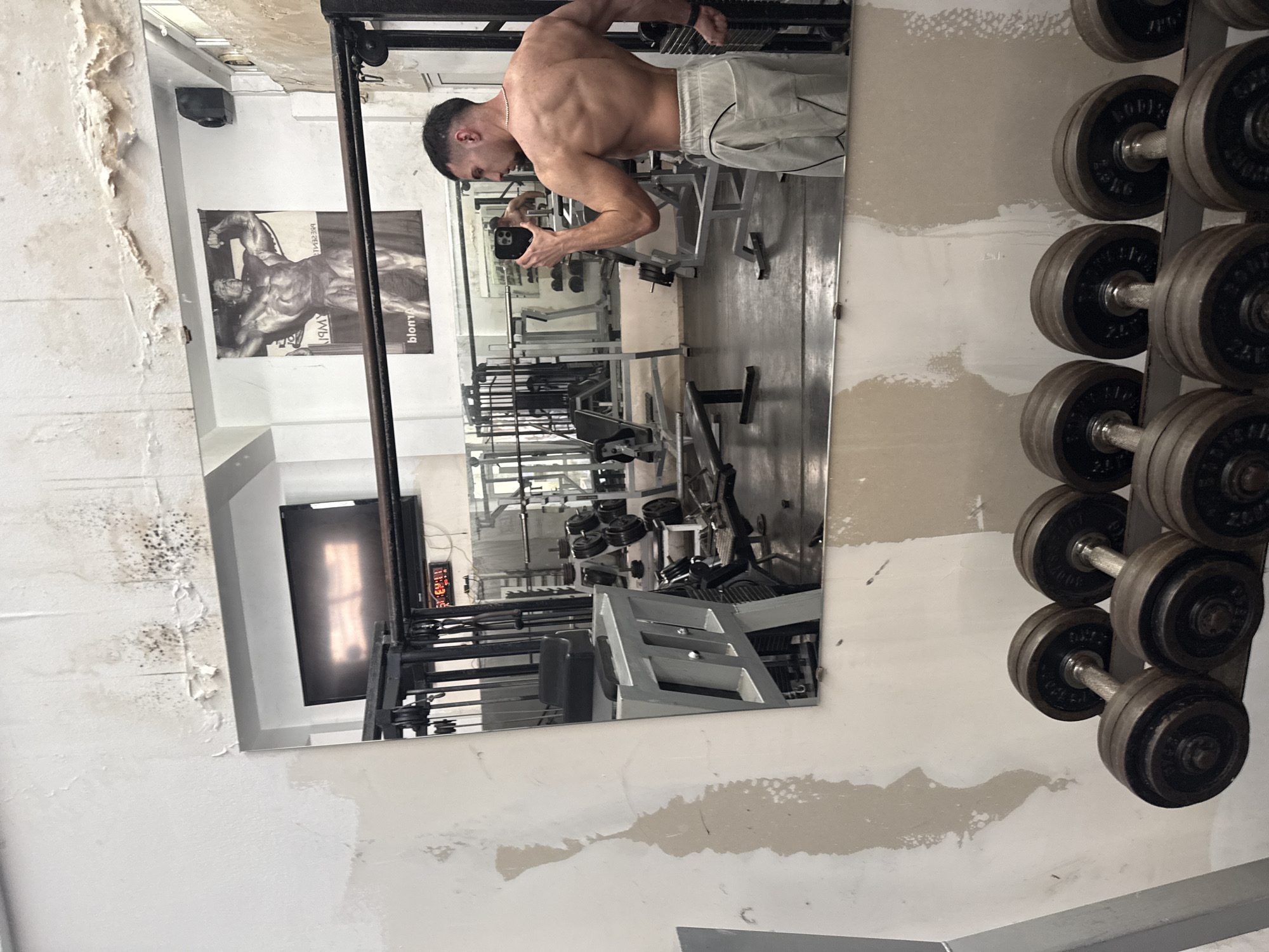 A muscular man with short dark hair and a fade haircut is depicted from the waist up, shirtless, and wearing light-colored athletic shorts, captured in a mirror selfie. He is flexing his back and arm muscles, holding a black smartphone to take the picture.
The setting is an indoor gym, possibly an older establishment, characterized by its worn appearance. The walls, painted white, show significant signs of wear and tear, including peeling paint, cracks, and dark patches resembling dirt or mold, particularly in the upper left section of the image.
Reflected in the large mirror are various pieces of gym equipment, such as weight machines (including cable machines and a leg press), benches, and weight plates. To the right of the mirror, a rack of dumbbells is visible, with plates marked "10 KG" and "20 KG" on some of them. Above a reflected black and white poster of a male bodybuilder, a black speaker is mounted on the wall. A television or monitor screen is also reflected in the lower left portion of the mirror, and a digital clock displaying "11:23" is visible in the reflection.
The man is engaged in the activity of documenting his physique, likely after a workout, through a mirror selfie. The poster text is partially visible as "BLOM..." and "BY AMB...".