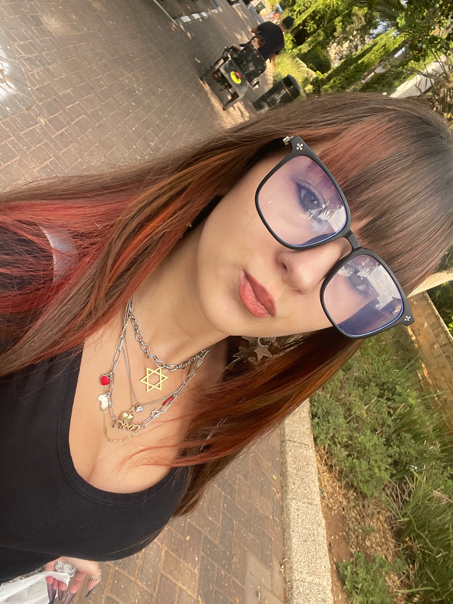 The image shows a selfie taken outdoors, with a young person wearing glasses and multiple necklaces. The background suggests a public space with paving stones, greenery, and a person in a wheelchair in the distance. The lighting indicates it is daytime, likely sunny. The environment appears to be a street or park area in Raanana, Israel, given the context. There are no specific landmarks visible to confirm the exact location within the city.