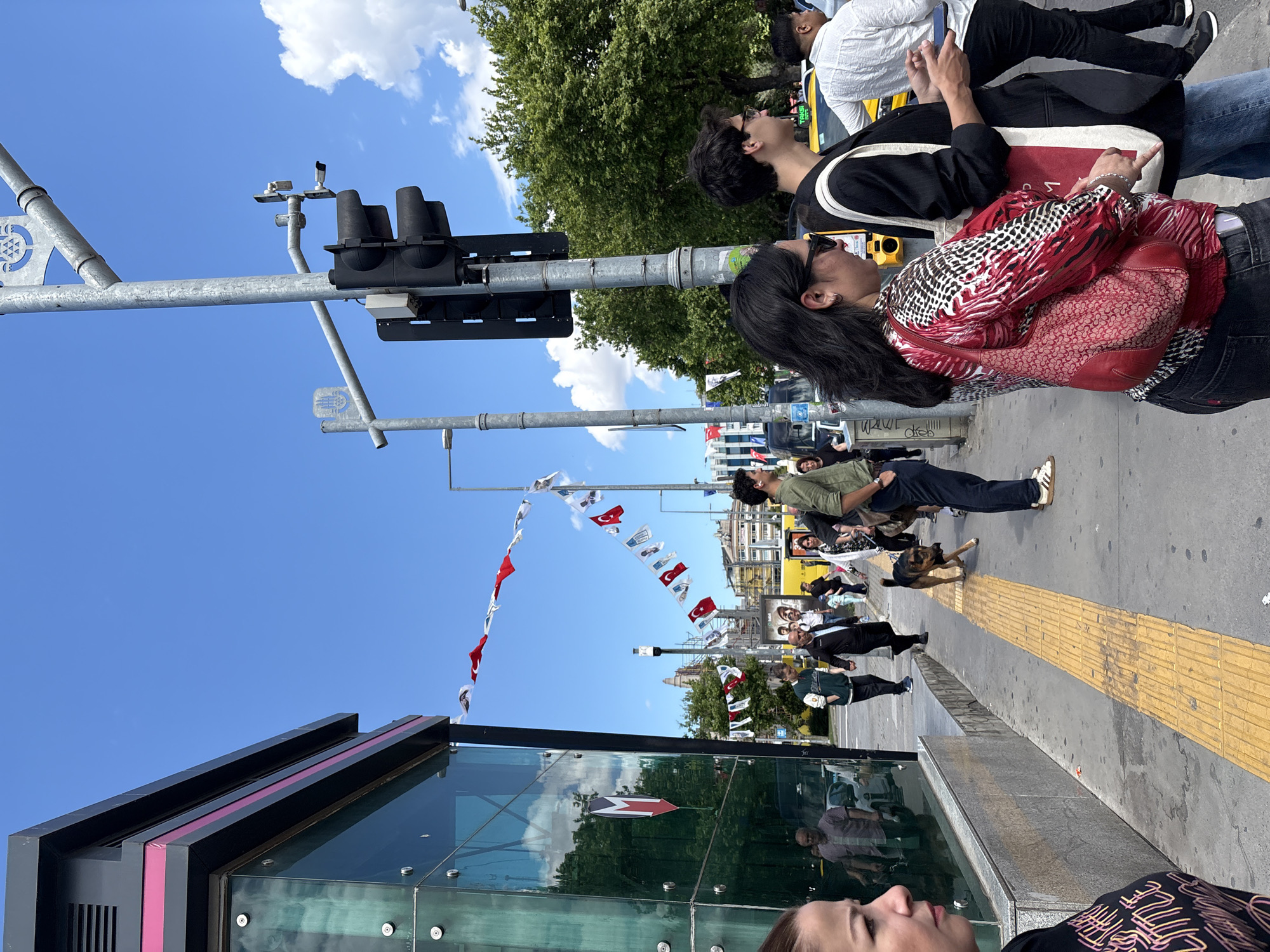 The image captures a street scene in Kadıköy, Turkey. The foreground features a modern bus stop with glass panels and a dark frame. A young woman with brown hair is visible in the bottom foreground. A traffic light stands prominently next to the bus stop, with a clear blue sky and some clouds overhead.
A string of Turkish flags hangs across the street, adding a festive touch. A line of people are walking along the sidewalk, some wearing sunglasses and looking upwards. A dog is walking on the sidewalk. Behind them, there is a yellow taxi cab.
The environment includes buildings and trees in the background, providing context for the location being an urban area. The overall scene depicts a lively street view in Kadıköy on a sunny day.