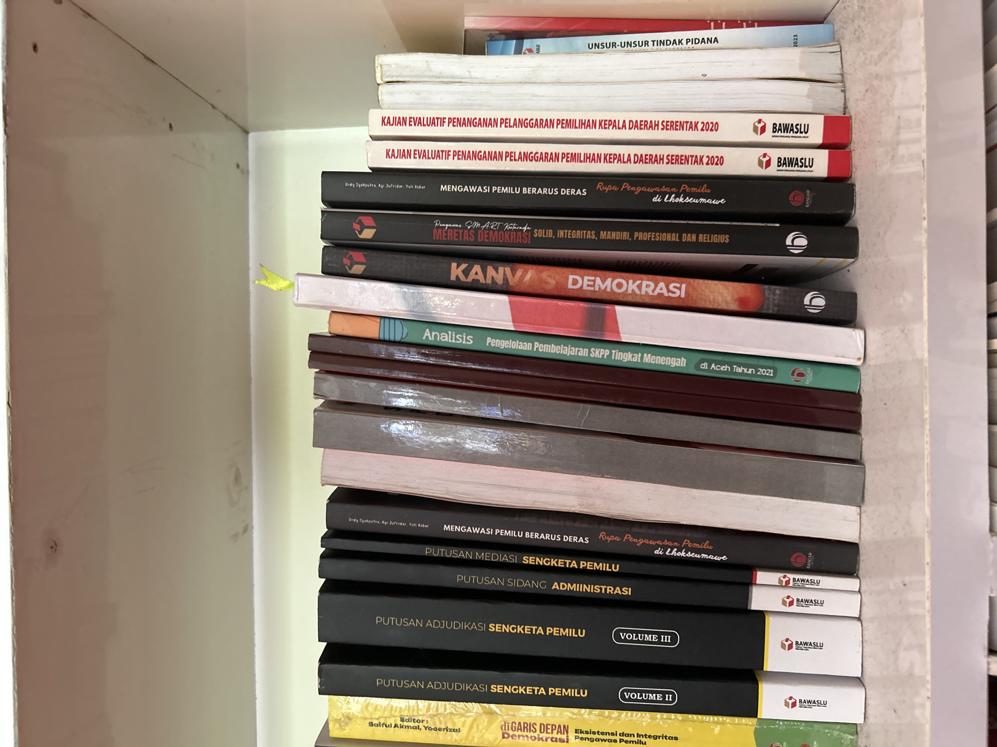 A stack of books is visible on a light-colored wooden shelf in an indoor setting. The books are mostly oriented with their spines facing outwards, displaying titles in Indonesian. Many of the books pertain to elections, democracy, and legal proceedings.
Visible text on the spines includes: "UNSUR-UNSUR TINDAK PIDANA" (Elements of Criminal Acts), with the year "2023" on an adjacent book. Several books are titled "KAJIAN EVALUATIF PENANGANAN PELANGGARAN PEMILIHAN KEPALA DAERAH SERENTAK 2020" (Evaluative Study on Handling Violations of Simultaneous Regional Head Elections 2020), bearing the logo of "BAWASLU" (Badan Pengawas Pemilu - Election Supervisory Agency).
Two books share the title "MENGAWASI PEMILU BERARUS DERAS Rupa Pengawasan Pemilu di Lhokseumawe" (Overseeing Elections with Strong Currents: Forms of Election Supervision in Lhokseumawe), authored by Drdy Syahputra, Agi Jufridar, and Yuli Asbar, directly linking to the provided location context. Other titles include "MERETAS DEMOKRASI SOLID, INTEGRITAS, MANDIRI, PROFESIONAL DAN RELIGIUS," "KANVAS DEMOKRASI" (Canvas of Democracy), and "Analisis Pengelolaan Pembelajaran SKPP Tingkat Menengah di Aceh Tahun 2021."
Further down the stack, titles related to legal aspects of elections are visible: "PUTUSAN MEDIASI SENGKETA PEMILU BAWASLU" (Mediation Decisions on Election Disputes BAWASLU), "PUTUSAN SIDANG ADMINISTRASI BAWASLU" (Administrative Hearing Decisions BAWASLU), and "PUTUSAN ADJUDIKASI SENGKETA PEMILU VOLUME III BAWASLU" and "VOLUME II BAWASLU" (Adjudication Decisions on Election Disputes Volume III and II BAWASLU). The bottom visible book is titled "di GARIS DEPAN Eksistensi dan Integritas Demokrasi Pengawas Pemilu" (At the Forefront: Existence and Integrity of Democracy Election Supervisors). The books suggest a collection focused on electoral law, observation, and democratic processes, particularly in Indonesia.
