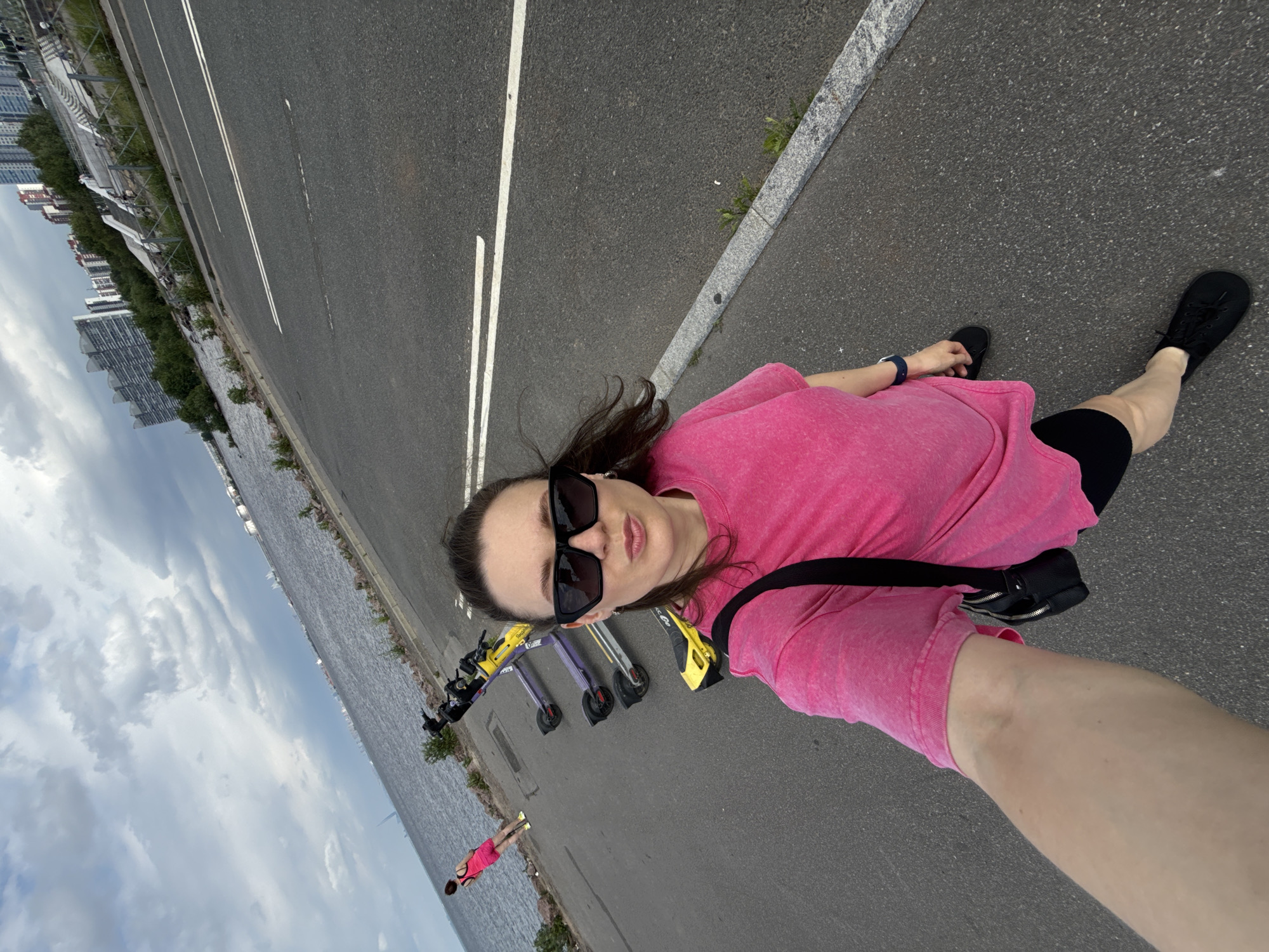 In Saint Petersburg, Russia, a woman in a bright pink t-shirt and black shorts takes a selfie on a paved pathway near a body of water. She is wearing black sunglasses and a black crossbody bag. Behind her, a line of parked electric scooters, some yellow and some purple, are visible. Further in the background, a woman in a pink top and shorts walks away from the camera towards the water. The scene is set against a cloudy sky and a cityscape with tall buildings. The pathway has white lane markings and a granite curb.