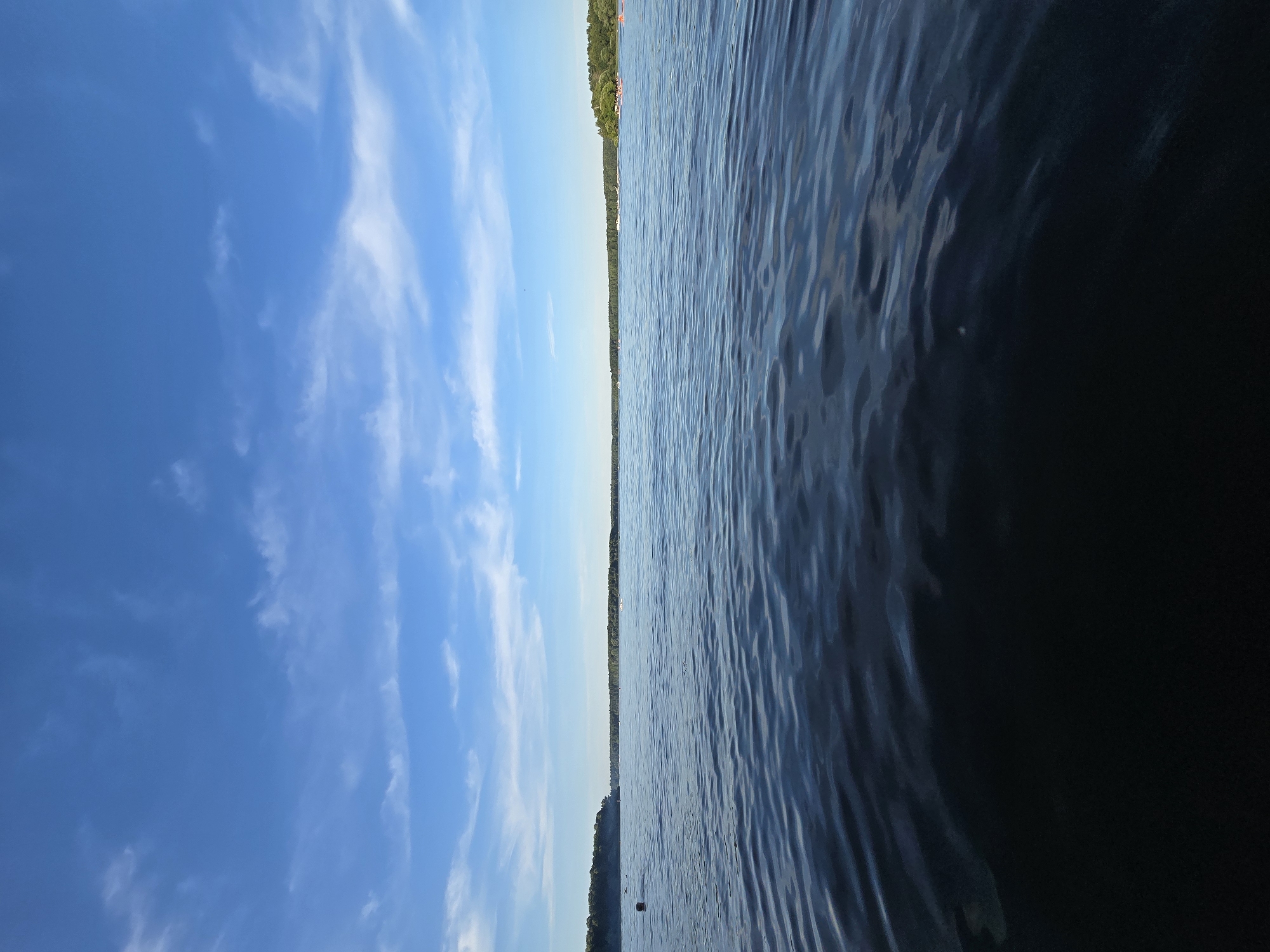 The image shows a wide expanse of dark blue water under a bright blue sky with scattered wispy white clouds. The foreground is dominated by the water, which has gentle ripples. In the mid-ground, a treeline of dense green foliage lines the distant shore. There are a few small boats visible on the water near the shore, and what appear to be buoys or markers closer to the left side of the frame. The scene appears to be a natural landscape, possibly a lake or a wide river, with a populated shoreline. The location is identified as посёлок гидроузла имени Куйбышева, Russia.