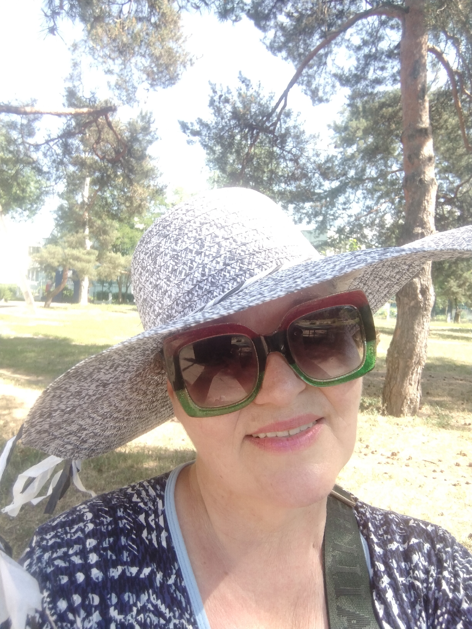 In a close-up, outdoor shot taken in Horishni Plavni, Ukraine, a woman wearing a wide-brimmed hat and large sunglasses smiles at the camera. She is wearing a patterned shirt. The background is blurred, showing trees and a grassy area, indicative of a park or outdoor recreational space. The lighting suggests it is daytime, likely sunny.