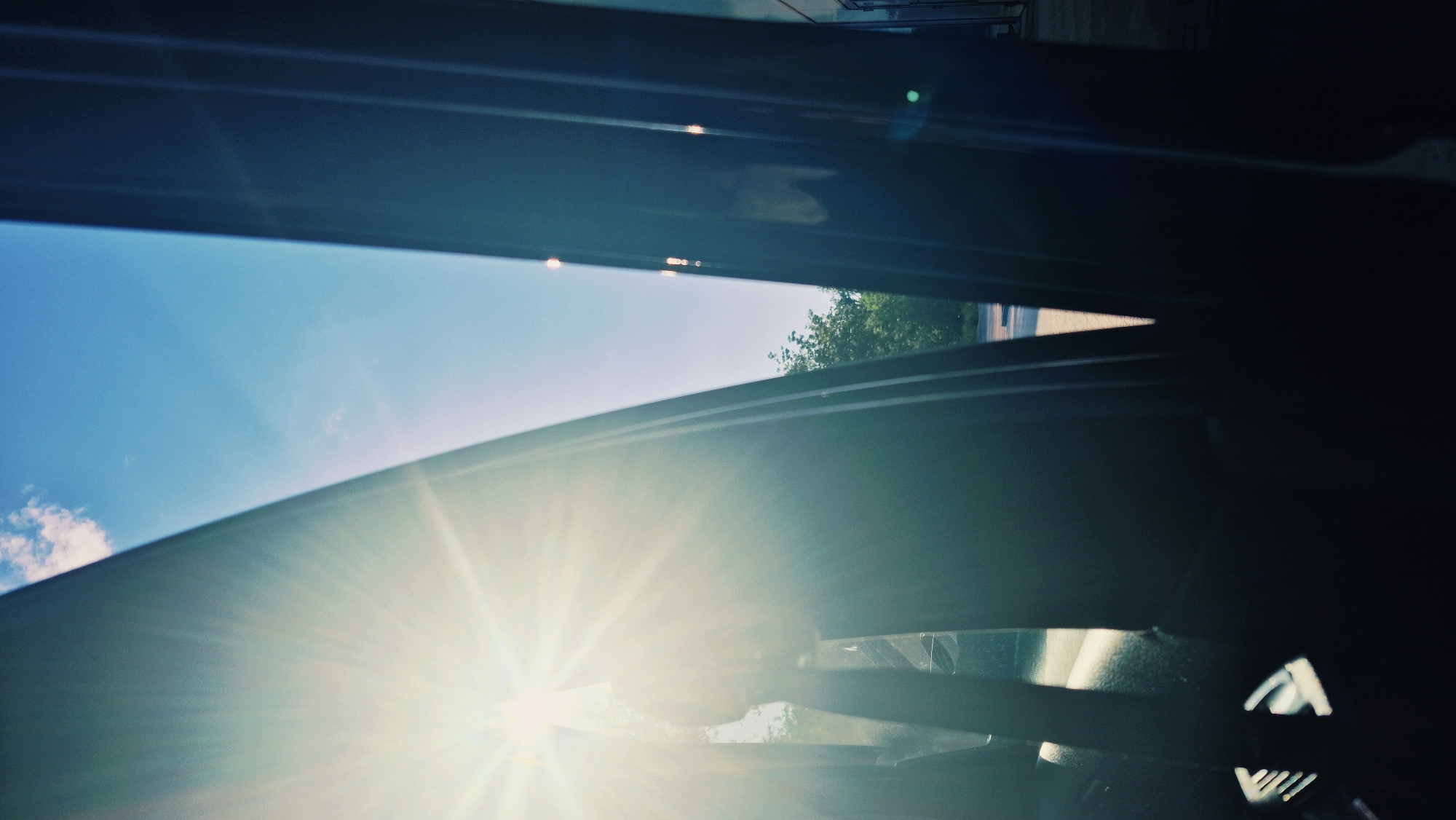 The image is an interior view from what appears to be the back seat of a car, looking outwards through an open door. Bright sunlight streams in from the left, creating a lens flare effect and illuminating a portion of the sky which is a clear, bright blue with a few small white clouds. The sun's rays are angled and prominent.
On the left side of the frame, the interior of the car is visible, including the dark fabric of a seat and the curve of a car door. Light is catching the edge of the car door, creating a bright, almost white line. On the right side, another car door or frame is visible, also dark, with vertical lines suggesting its structure.
Through the opening of the car doors, an outdoor scene is visible. There are green trees and foliage, suggesting a park or residential area. A paved surface, possibly a road or sidewalk, is also discernible. The light suggests it is daytime, likely mid-morning or afternoon.
The image has a slightly washed-out, vintage, or artistic quality due to the intense sunlight and potential filters or editing. There are no people clearly visible in the image.
Based on the visual cues alone, it is not possible to definitively determine the location within Chișinău, Moldova.
