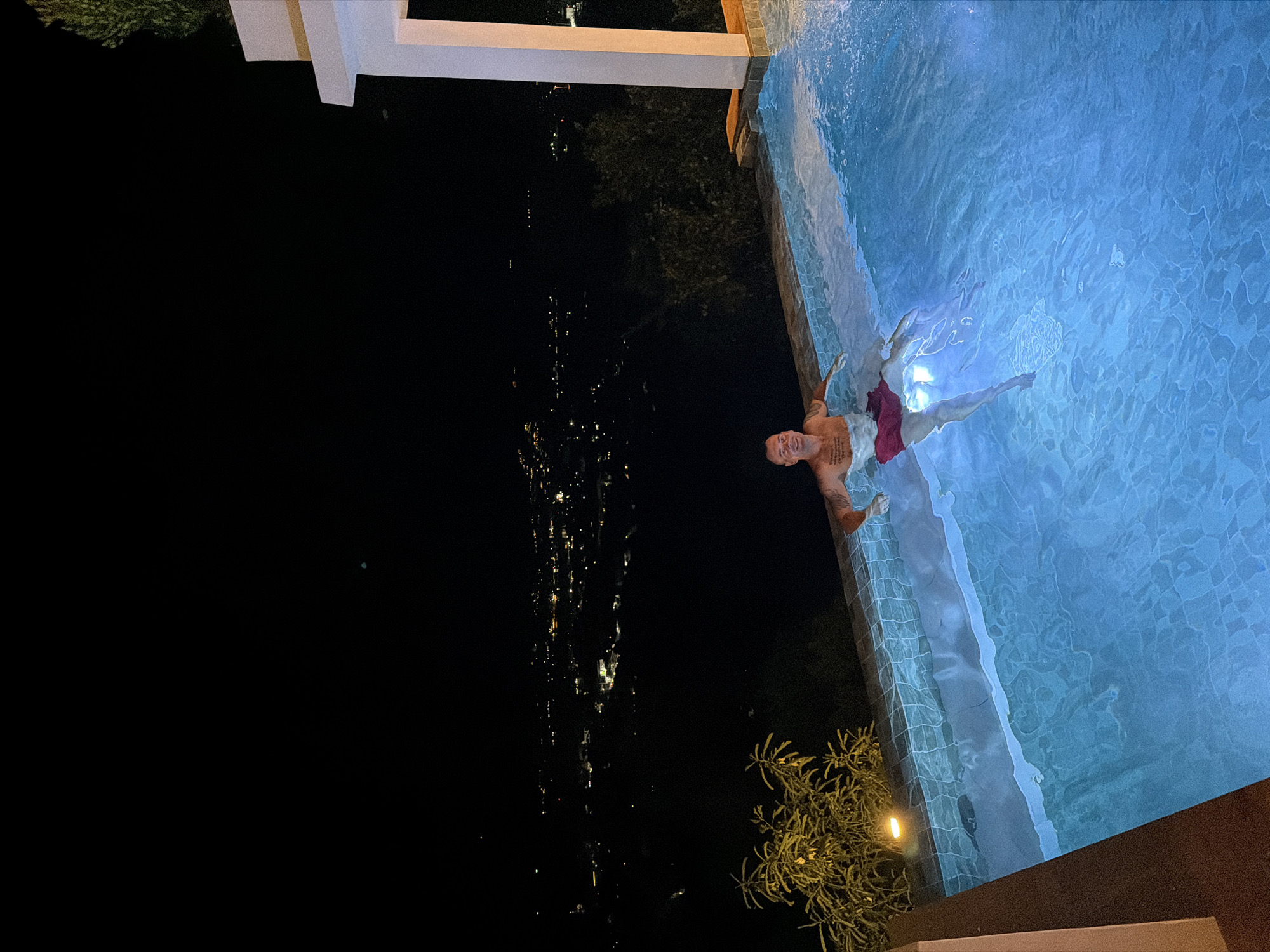 A man is in a swimming pool at night in บ้านบางรักษ์, Thailand. The pool is lit with blue light, illuminating the water. The man is wearing red shorts and floating on his back. In the background, city lights are visible in the distance, stretching out across the night sky. A white building structure is partially visible above the pool. A bush or shrub with yellow lighting is visible to the bottom right of the pool.