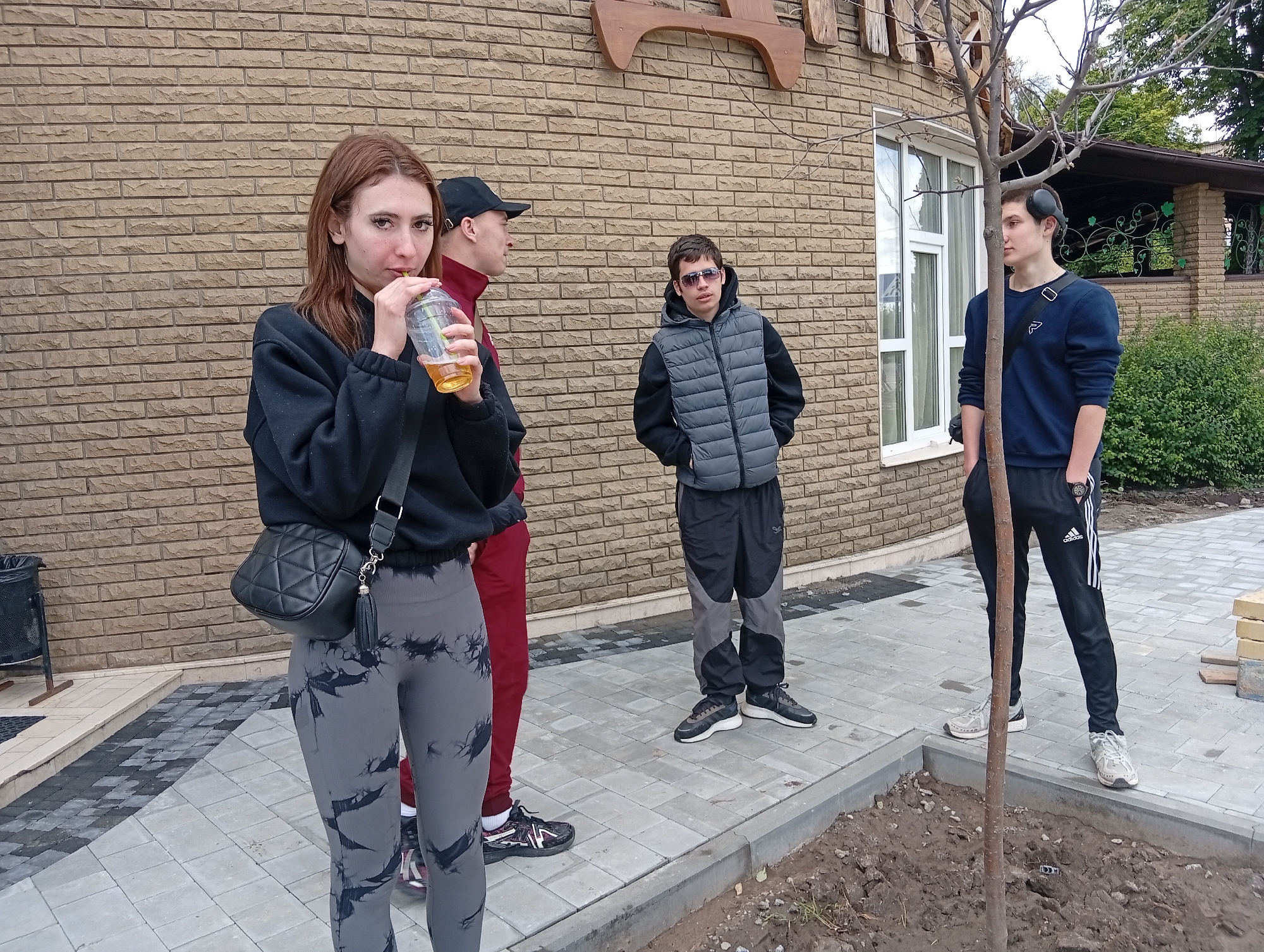 The image shows four people standing on a paved area in front of a brick building. A young woman in gray leggings and a black sweatshirt drinks from a clear plastic cup. Next to her, a person in a red tracksuit and a black cap is partially visible. A young man wearing sunglasses and a gray puffer vest stands with his hands in his pockets. To the right, a young man in a blue sweatshirt and Adidas pants stands near a newly planted tree. The location appears to be a building exterior, likely in Poltava, Ukraine.