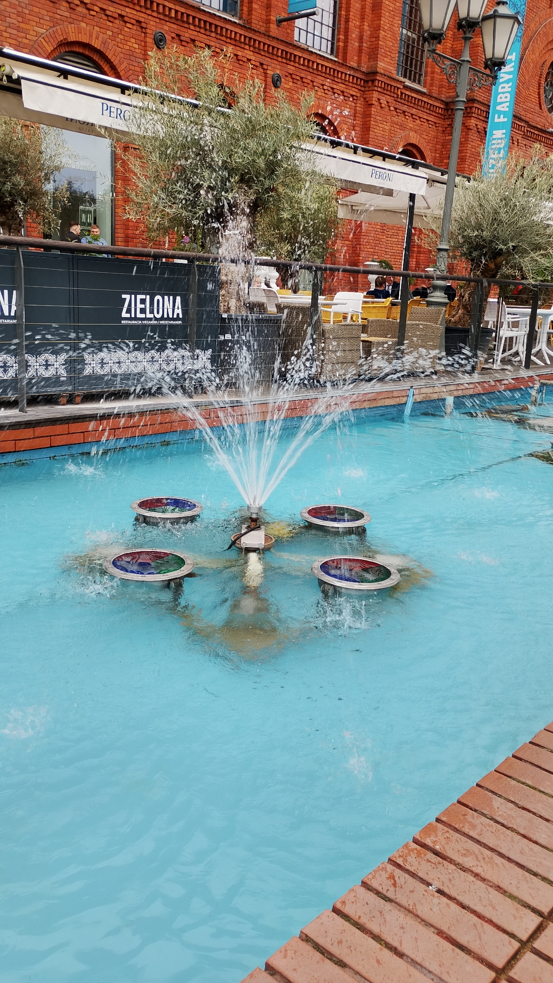 The image shows a water fountain in the Manufaktura complex in Łódź, Poland. The fountain features a central nozzle shooting water upwards, surrounded by four circular floating objects with colored sections. The water in the fountain is a light blue hue. Brickwork lines the edge of the fountain. In the background, there is a brick building with a restaurant named "Zielona" and umbrellas branded "Peroni". There are trees and a lamp post visible. There are people at tables in the background, partially obscured by the fountain.