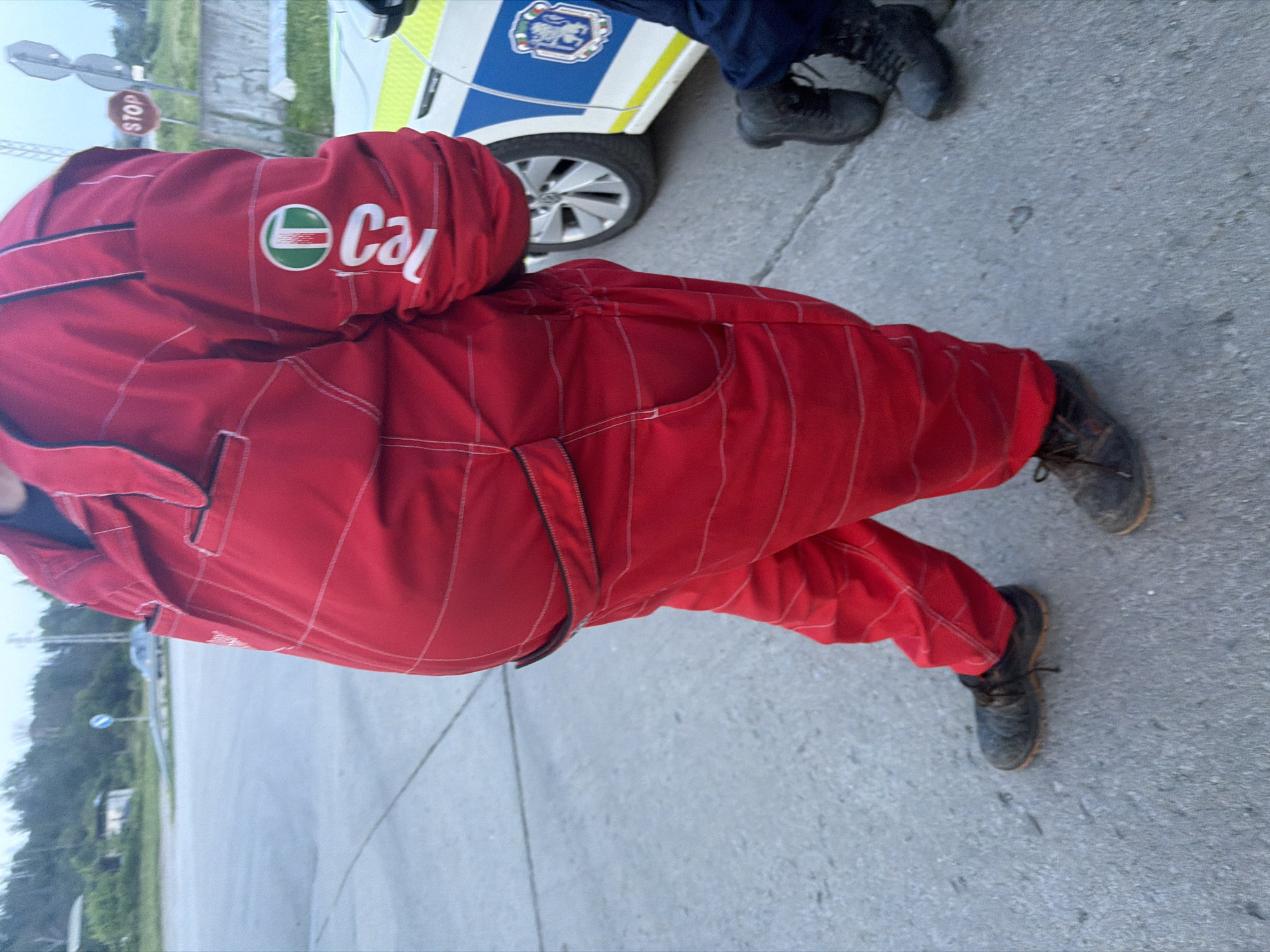 The scene is set in Veliki Preslav, Bulgaria, under overcast daylight conditions, on an asphalt or concrete surface. In the foreground, a large individual, partially cropped, stands facing slightly left of the camera. This individual is wearing a full-body red overall or racing suit, featuring a black collar and black zippered pockets on the chest and waist. White stitching forms a grid pattern across the suit. On the right sleeve, a vertical white "Castrol" logo is present, topped by a circular green Castrol emblem with white and red internal elements. The person wears dark, worn lace-up shoes with light-brown soles. To the right, partially visible, is the side of a stationary white police vehicle, identifiable by a yellow stripe along its lower side and a blue section on the rear door bearing an official emblem depicting a shield with a lion rampant, characteristic of Bulgarian police. A front wheel of this vehicle is visible. Adjacent to the police vehicle, the lower legs and feet of a second individual, identified as a police officer, are visible, clad in dark blue trousers and dark boots. In the background, a paved road or open area extends, bordered by dense trees and foliage. A blue circular road sign is visible on the left side of the background, and on the right, a red octagonal "STOP" sign is visible alongside another partially obscured sign, situated near a concrete barrier.
