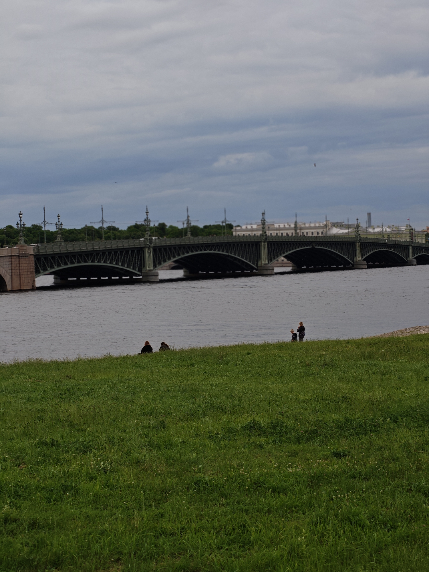 The image depicts a wide river in Saint Petersburg, Russia, spanned by a grand, arched bridge. On the grassy bank in the foreground, two individuals are seated, facing away from the camera, while further along the bank, two more individuals, one appearing to be an adult and the other a child, stand facing the water. The sky is overcast with dramatic clouds, and the water reflects the subdued light. The bridge, a prominent architectural feature, shows intricate metalwork and stonework. The scene is peaceful, with the figures on the bank appearing contemplative or enjoying the view.