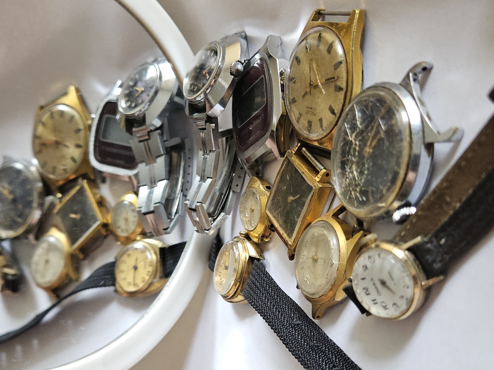 The image shows a collection of vintage wristwatches, likely a personal collection, displayed against a white background that appears to be a shelf or display case. The scene is brightly lit, suggesting an indoor setting. The watches are varied in style, with several gold-toned cases and metal bands, some with leather straps. One watch prominently features the text "ПОЛЕТ" which is a Russian watch brand. Another watch has text that appears to be "ПОБЕДА" and "СДЕЛАНО В СССР" (Made in USSR). The watches are arranged in a somewhat haphazard manner, overlapping each other, creating a sense of abundance and history. The overall impression is one of preserved timepieces, perhaps awaiting sale or being admired for their vintage appeal. The location context of Ulyanovsk, Russia, is relevant as it aligns with the presence of Soviet-era watches like Poljot and Pobeda. There is no indication of a specific time of day or weather conditions, and no people are visible. The emotion conveyed is primarily one of nostalgia and appreciation for craftsmanship.