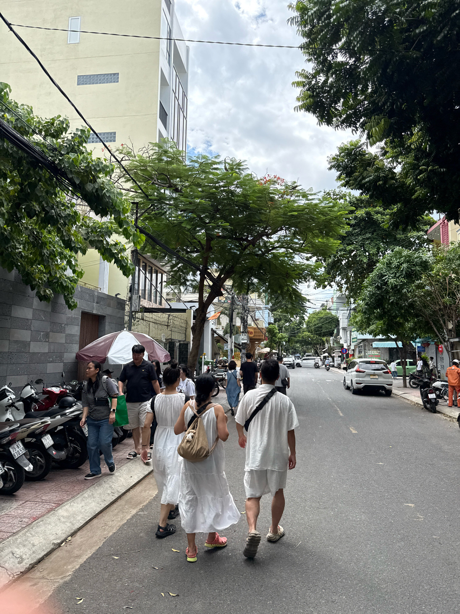 In Nha Trang, Vietnam, a street scene unfolds with a group of people walking away from the camera on a gray asphalt road. The street is lined with buildings of varying architectural styles, some with balconies and windows, and others featuring stone facades. Lush green trees with dense foliage overhang the street, casting shadows. Several parked motorcycles are visible on the left side of the road, some with vibrant red accents. A white and brown umbrella is set up on the sidewalk. In the distance, traffic, including cars and motorcycles, moves along the street under a cloudy sky. A person in an orange uniform stands on the right side of the road. The overall impression is a bustling urban environment during daylight hours.