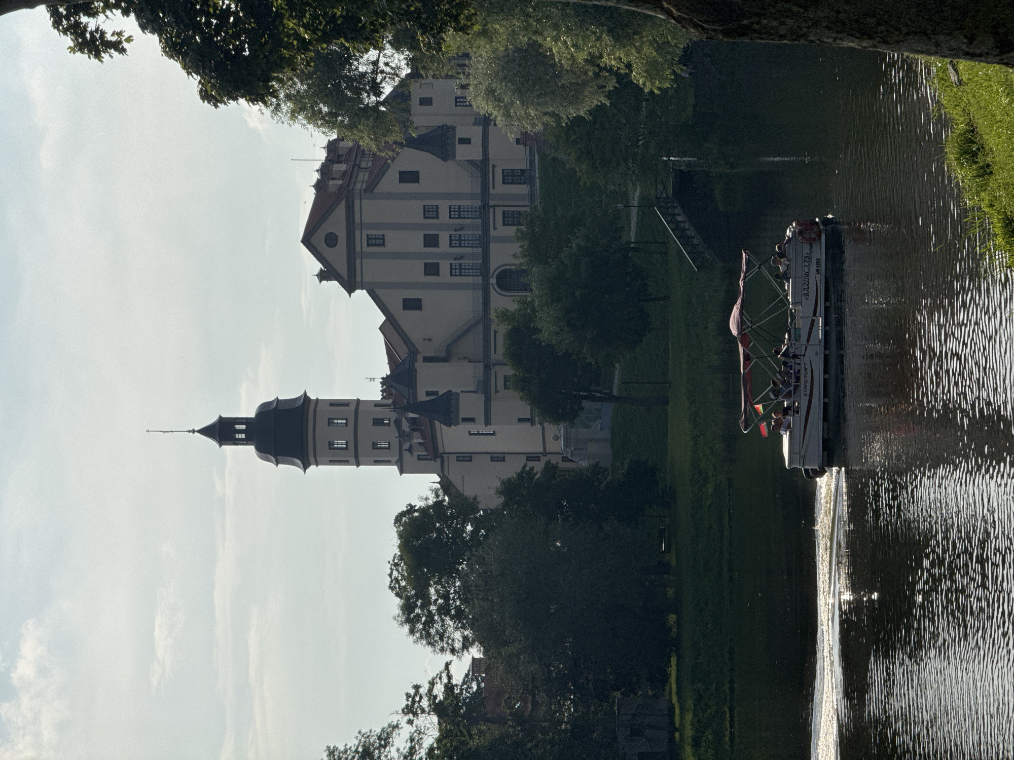 The image shows a wide shot of the Nesvizh Castle in Nesvizh, Belarus, on a partly cloudy day. The castle, with its distinctive tower, is situated on a gentle incline, surrounded by lush green trees and a tranquil body of water in the foreground. A white boat, carrying several passengers, is cruising on the water, creating ripples that catch the sunlight. The boat has a red canopy and appears to be on a tour. In the background, a wooden bridge can be seen arching over a waterway, suggesting a park-like setting around the castle. The overall atmosphere is serene and picturesque, capturing a moment of leisure against the backdrop of historical architecture.