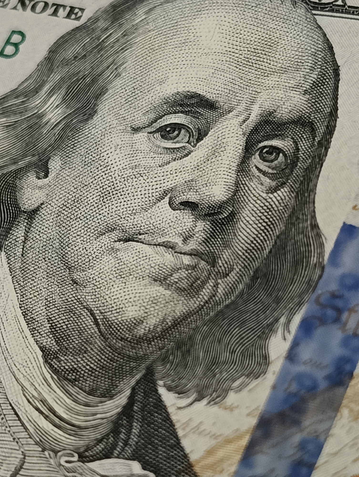 A close-up shot shows Benjamin Franklin's face on a United States one-hundred dollar bill. The image focuses on his eyes, nose, and mouth, highlighting the intricate details of the engraving. The bill is partially obscured by a blue security ribbon.
