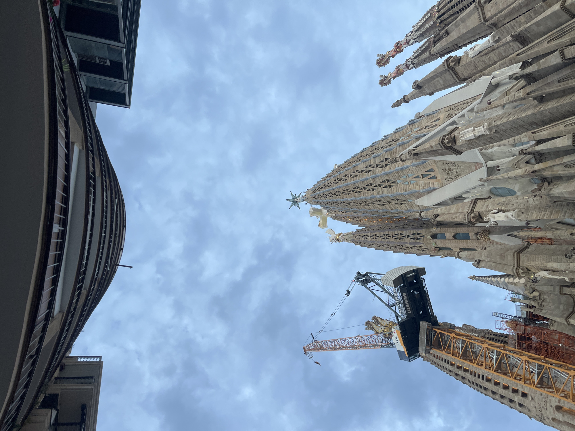 The image captures a low-angle perspective of the Sagrada Familia basilica in Barcelona, Spain, under a cloudy sky. A large construction crane, predominantly orange with a black and white cab, is positioned to the left of the basilica's spires. The basilica itself, made of intricate stone, displays several spires, one of which is topped with a star. The building's facade is richly detailed with sculpted elements. In the upper portion of the frame, the curved balconies of a modern building are visible, contrasting with the historic architecture of the Sagrada Familia. The scene suggests ongoing construction or renovation work at the iconic landmark.