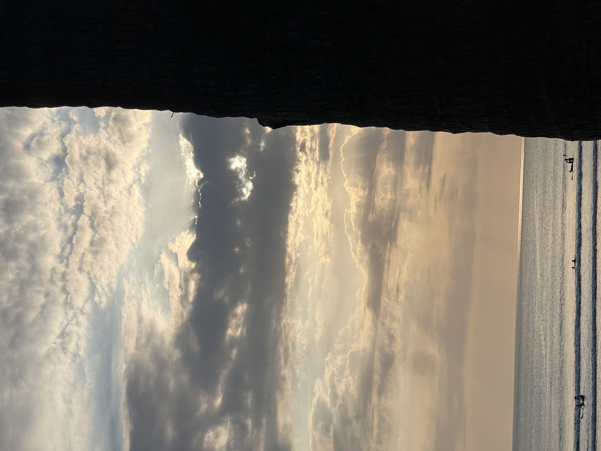 The image depicts a scenic view of the ocean at Kuta, Indonesia, during what appears to be sunset or sunrise. A large, dark, textured object, likely a palm tree trunk, dominates the right side of the frame, casting a shadow over the scene. The sky is filled with a dramatic display of clouds, with shafts of light breaking through, illuminating patches of the sky and the ocean surface. The sea is calm with gentle waves rolling in. In the distance, several individuals are visible in the water, some on surfboards, suggesting a surfing activity. The overall atmosphere is serene and captures the natural beauty of Kuta's coastline.