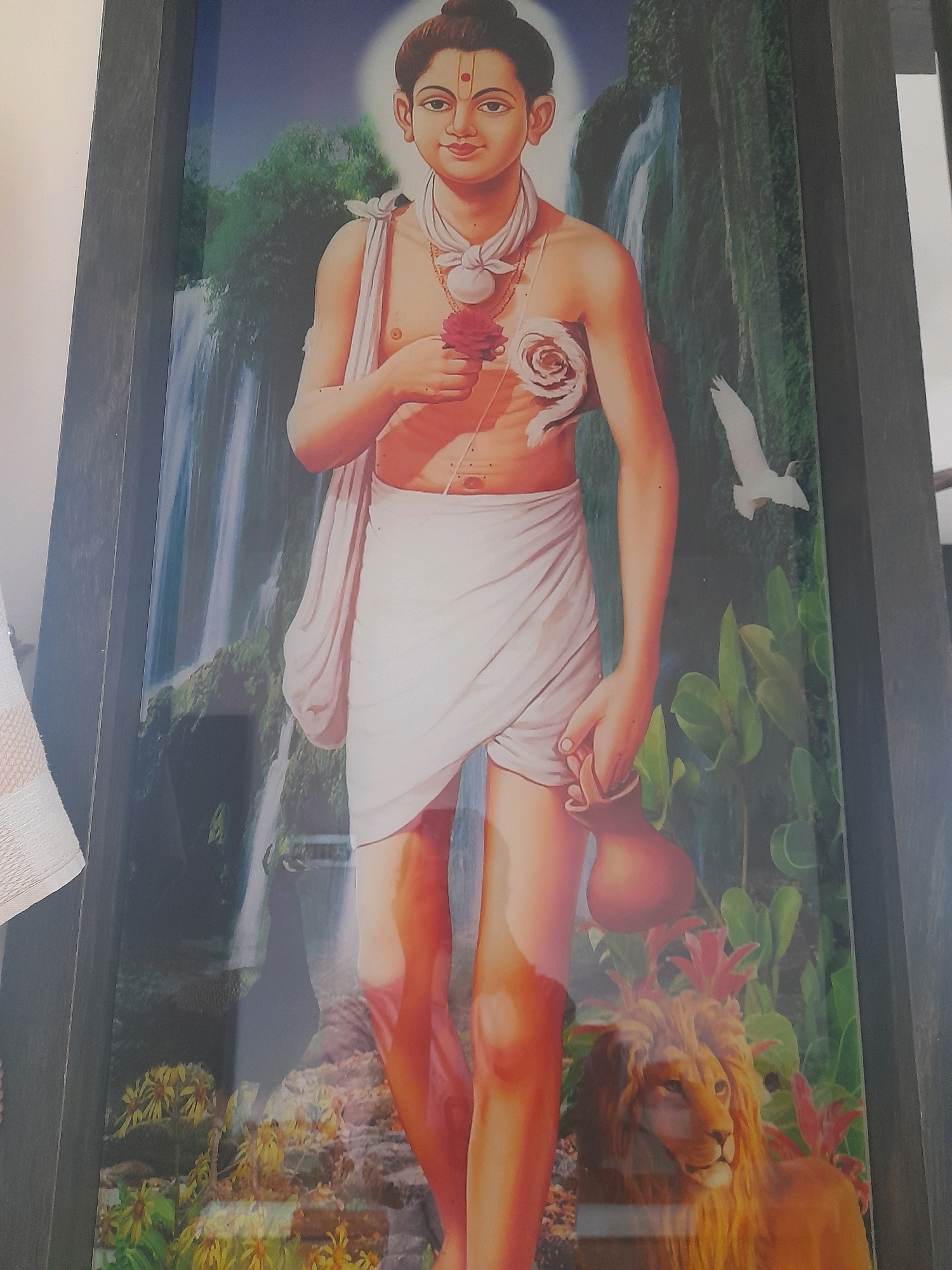 A full-body portrait of a young male figure, depicted in a realistic style, stands centrally within a framed artwork. The figure has dark hair pulled back and wears a red bindi on his forehead. He is adorned with a white cloth draped around his waist and a light-colored sash across his chest, from which hangs a necklace with a floral pendant. In his right hand, he holds a red rose, and his left hand clutches a small, brown pitcher. His chest is bare, revealing decorative floral markings.
The background is a vibrant, natural landscape featuring lush green foliage and a waterfall cascading down rocky cliffs. To the right of the figure, a white bird is depicted in flight, and below, a majestic lion with a golden mane rests among the plants. The bottom portion of the artwork displays a cluster of yellow flowers and rocky terrain. The overall impression is one of serene power and natural beauty. The artwork is encased in a dark, wooden frame, and the image appears to be a photograph of this framed artwork.