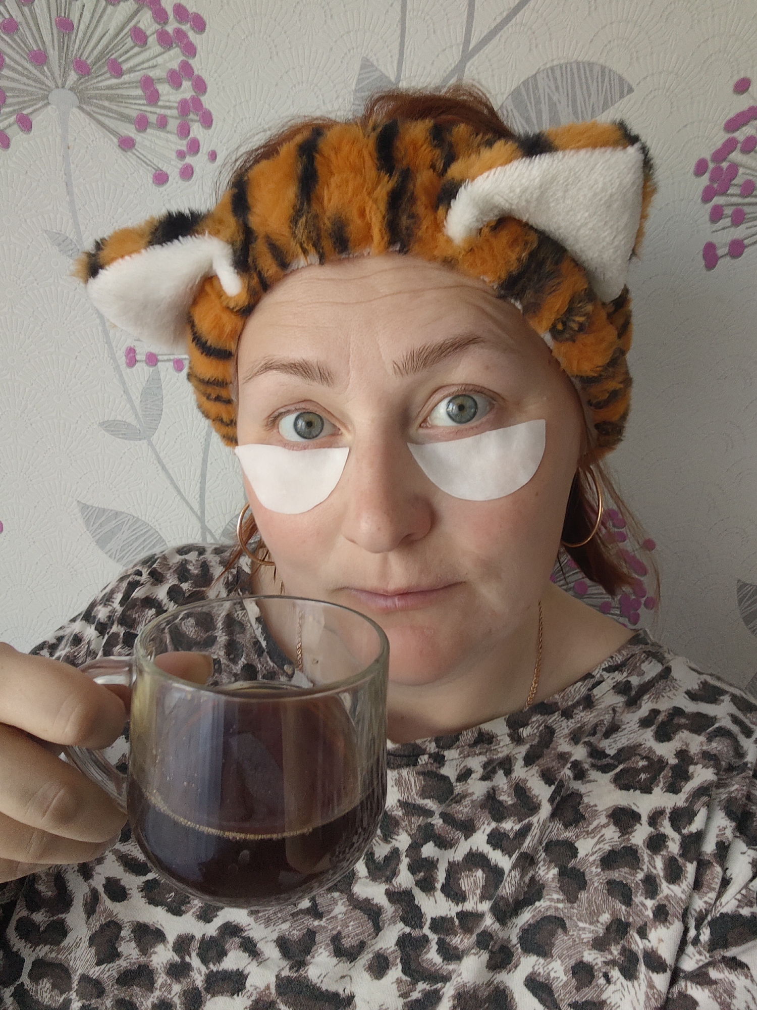 A woman with fair skin, wearing a tiger-striped headband with ears and white under-eye patches, holds a clear glass mug filled with dark liquid, likely coffee. She is wearing a leopard print shirt. She has light green eyes and light red hair. She is also wearing gold hoop earrings and a gold necklace. The background is a light gray wallpaper with a pattern of stylized gray and pink floral designs. The location is Markovskii, Russia.