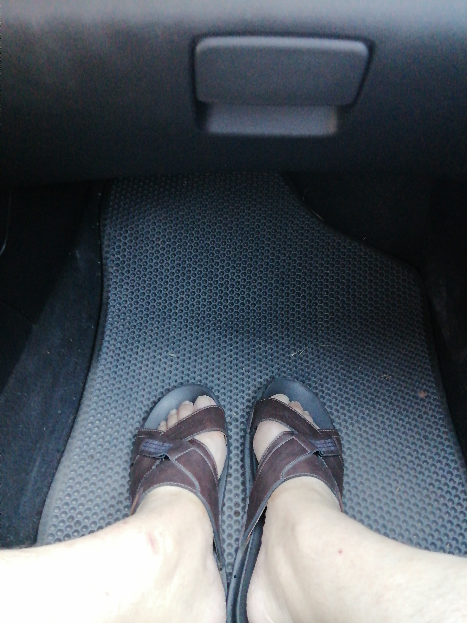 The image shows a first-person perspective of a person's feet wearing brown slide sandals, positioned on the floor of a vehicle. The floor is covered with a textured black mat. Above the mat is a dark grey interior surface, likely the dashboard or glove compartment area, with a black handle or latch visible. The scene appears to be indoors or in a sheltered environment, consistent with the interior of a car. The lighting suggests it is daytime. The context of Rostov-on-Don, Russia, is provided as a location clue, but there are no visible elements in the image that specifically indicate this location.