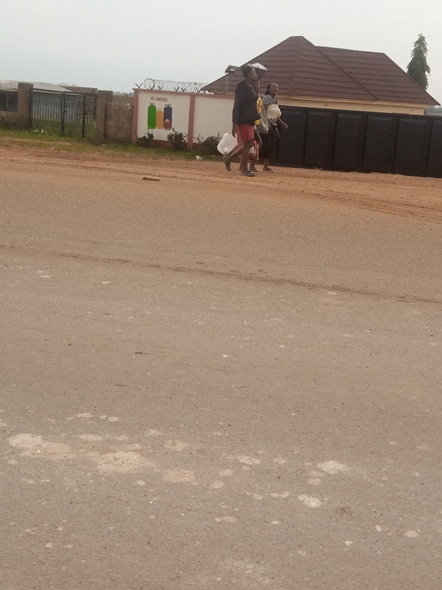 Three individuals are walking on a dirt road next to a paved road. A building with a brown tiled roof is visible behind them, with a black metal fence in front of it. A white wall with a "NO SMOKING" sign and images of gas cylinders is to the left of the building. The sky is overcast.