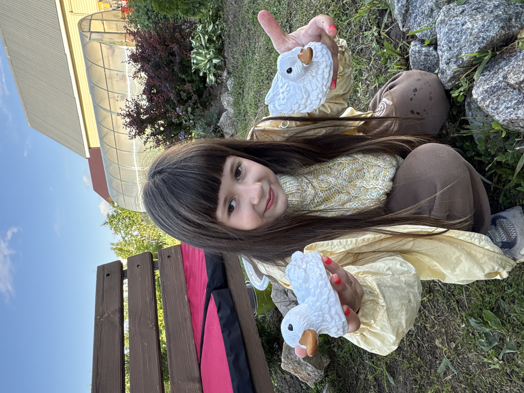 A young girl is captured in a cheerful outdoor portrait, smiling brightly at the camera, likely in Chaikovsky, Russia. She has long, dark hair with bangs and is wearing a cream-colored knitted sweater under a yellow outer garment, paired with brown pants. She holds up two identical white goose figurines with orange beaks, one in each hand. Her fingernails are painted red.
The setting appears to be a sunny garden or backyard. In the background, there is a yellow building with a gray corrugated roof, adjacent to a clear, tunnel-shaped greenhouse. Lush green grass, dark red foliage bushes, and several light-colored rocks are visible around her. A wooden fence or bench back is partially visible on the left side of the frame. The bright daylight and clear sky suggest pleasant weather. No visible text is present in the image.