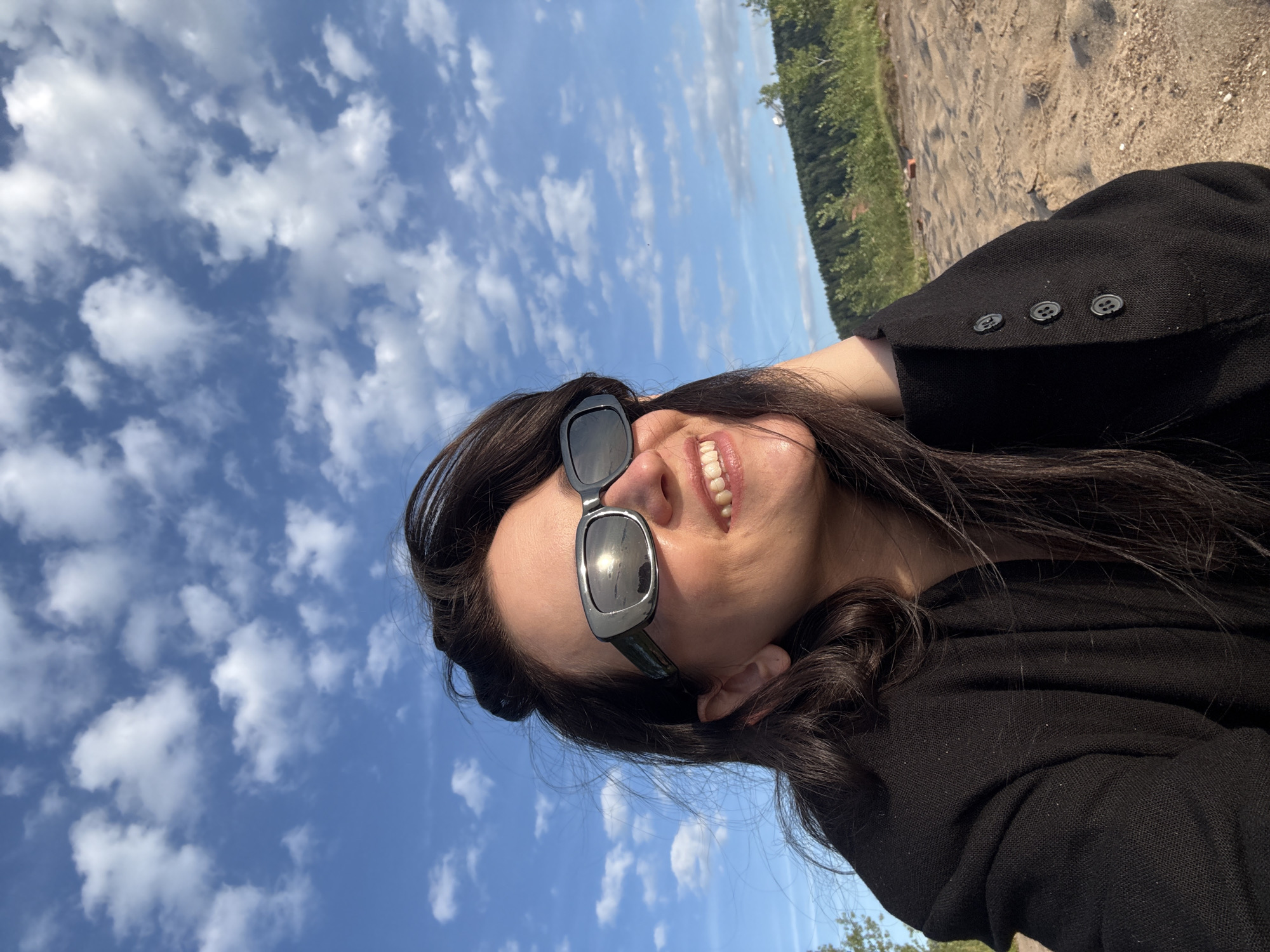 A woman is captured in a selfie outdoors, with a bright blue sky filled with scattered white clouds dominating the background. She is wearing large, dark sunglasses and a black jacket with three dark buttons visible on the right side of the frame. Her dark hair is slightly windswept. She is smiling, with her teeth showing, and her right hand is casually resting on the back of her head. The ground beneath her appears to be sand, and behind her, there is a line of green trees and vegetation on a gentle slope. The overall lighting suggests it is daytime, possibly late afternoon due to the warm tones. The location appears to be a natural setting, perhaps a beach or a sandy bank along a body of water, consistent with the waterfront areas of Chaykovsky, Russia.
