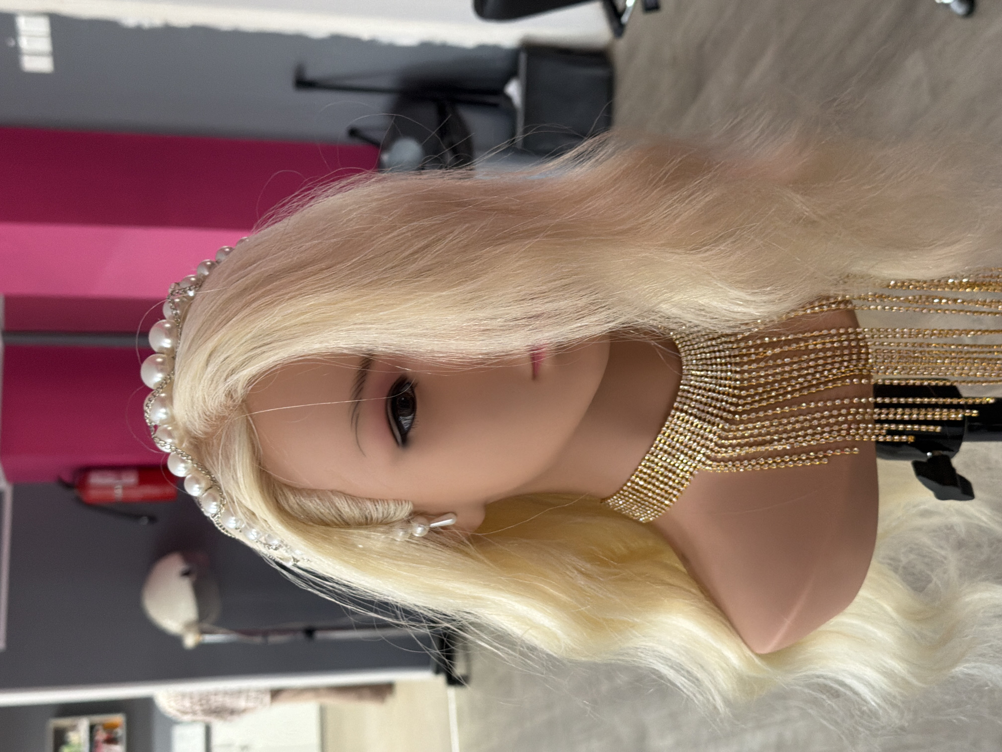 The image shows a blonde mannequin head with a pearl and rhinestone headband, pearl earrings, and a sparkly gold necklace. The mannequin is positioned in front of a wall that is partially pink and partially gray. In the background, there is a red fire extinguisher, a hair dryer on a stand, and shelves with items. The floor is a light wood laminate. The image appears to be taken indoors, possibly in a salon or beauty shop. There is no information in the image to indicate the location is Kyiv, Ukraine.