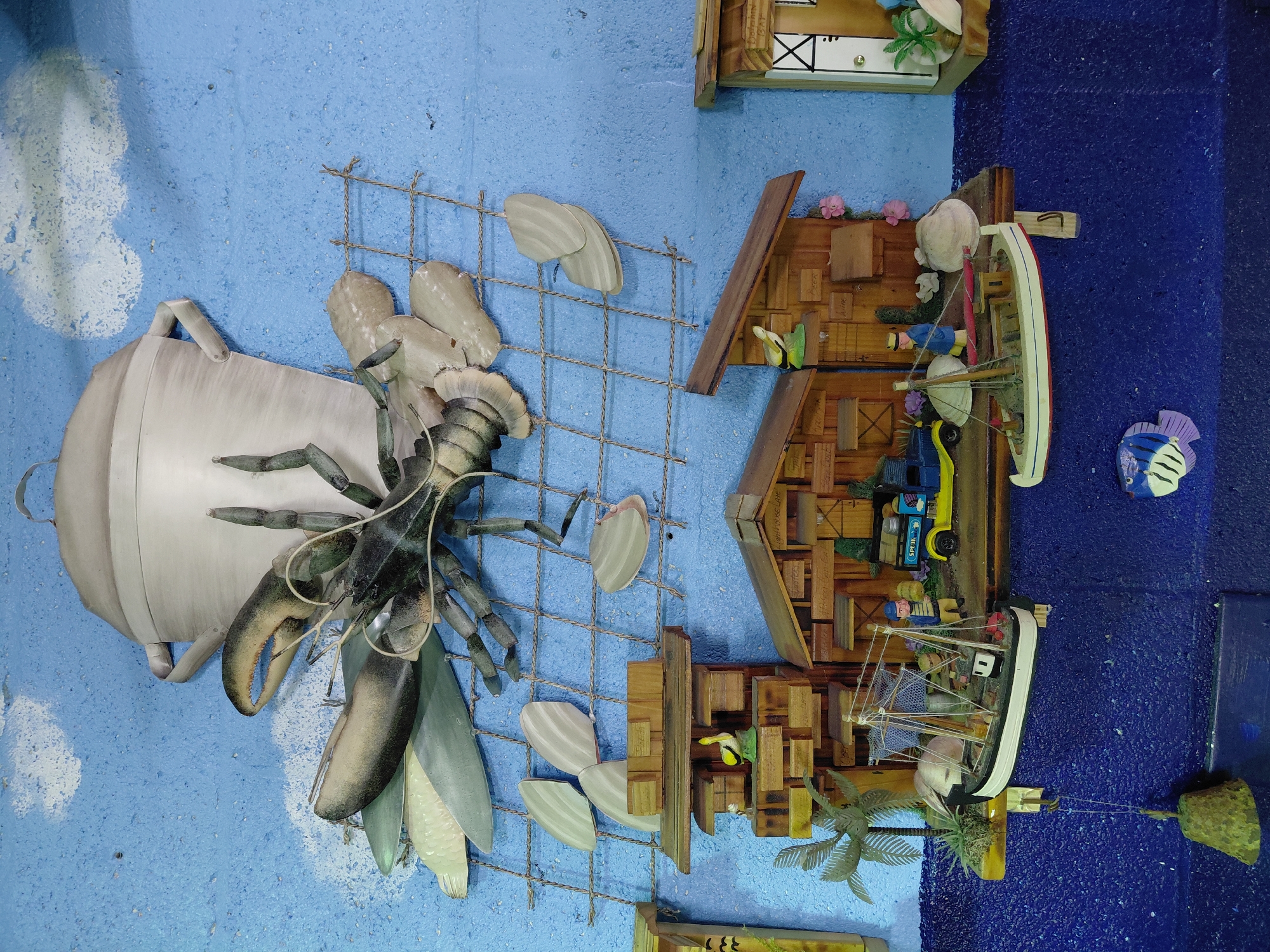 The image displays a nautical-themed wall decoration, likely within a home or establishment in Cutler Bay, Florida. The scene presents a blend of sculpted and crafted elements mounted on a blue-painted wall.
On the left, a large, sculptural representation of a lobster emerges from a pot. This assembly is connected to a wire mesh, supporting what appear to be stylized shell shapes.
Moving to the right, a miniature wooden diorama depicts coastal scenes. This includes stylized wooden houses, a pier with model boats, and miniature figures of people. The backdrop transitions to a darker blue, suggesting a deeper part of the water. A small decorative fish is also present.