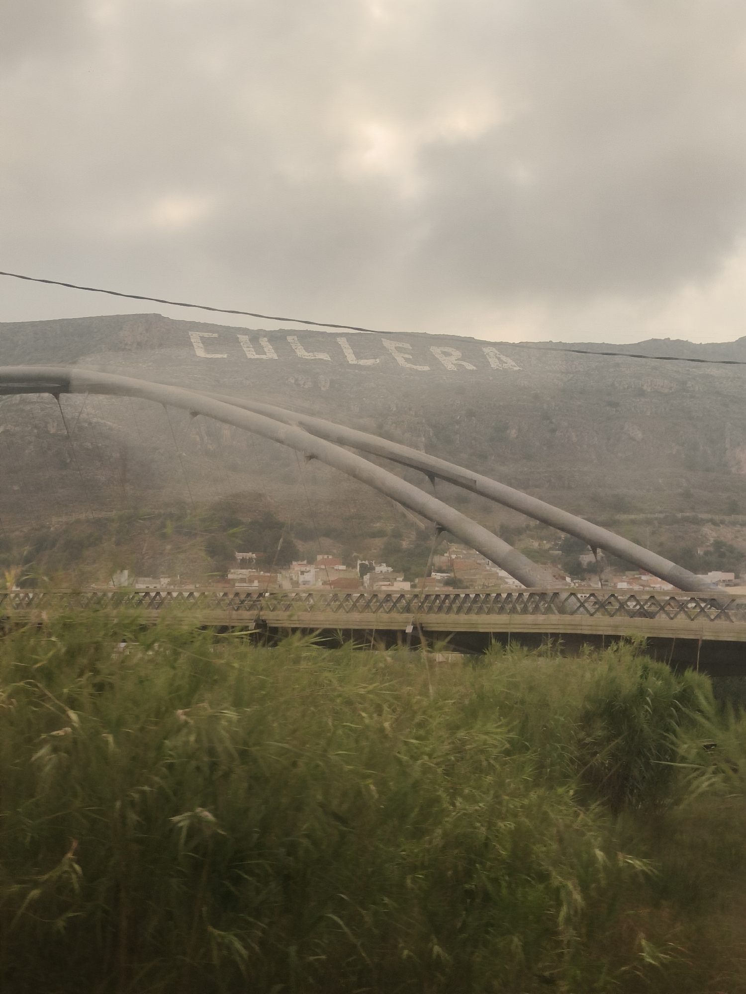 The image captures an overcast sky above a hillside where large, light-colored letters spelling "CULLERA" are visible. A modern bridge with distinctive arching supports spans across the middle ground. Below the bridge, a village with red-tiled roofs and white buildings is nestled against the hillside. In the foreground, dense green vegetation, possibly reeds or tall grass, obscures the lower portion of the scene, suggesting a viewpoint from ground level near a body of water or marshland. The overall impression is one of a somewhat hazy, possibly dusty, environment.