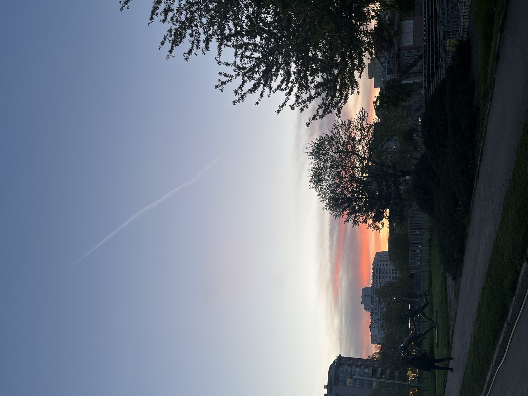 The image displays a serene evening or morning scene captured in Sofia, Bulgaria, characterized by a dramatic sky gradient. The upper portion of the frame is dominated by the sky, transitioning from a soft, light blue on the left to vibrant horizontal streaks of orange, pink, and red towards the right, indicative of either sunset or sunrise. A faint, white contrail extends diagonally across the lighter blue section of the sky.
Silhouetted against this colorful backdrop are numerous trees and foliage. A large, dense tree branch extends into the upper left, while another tree with intricate branches is positioned further right, both appearing dark against the bright sky. Along the right edge, dark bushes and a fence line are visible.
Below the horizon, various urban structures are present. Buildings with some illuminated windows are silhouetted against the colorful sky, suggesting a residential or commercial area. In the lower right, a paved pathway runs alongside a strip of green grass. Within this grassy area, several dark, human-like statues are distinctly visible, appearing to be in dynamic poses, perhaps dancing or engaged in an activity. A single person, also silhouetted, stands near these statues or further down the path.
The overall impression is one of peaceful urban nature at a transitional time of day, with the rich sky colors providing a striking contrast to the dark silhouettes of the foreground elements. There is no visible text in the image.