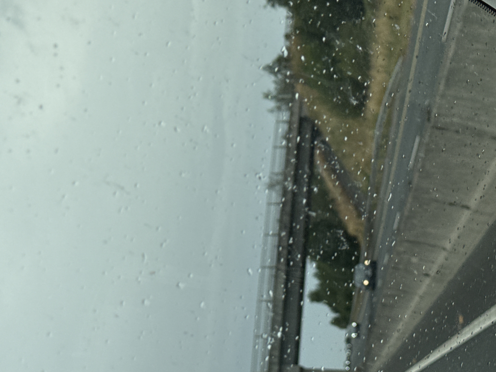 The image depicts a view from inside a vehicle driving on a wet highway in Craan, Ireland. Raindrops are visible on the windshield, obscuring some details of the scene. In the distance, a concrete overpass spans the highway, with trees and vegetation lining the sides. Several cars are driving on the highway, with headlights illuminated, suggesting low visibility due to the weather. The overall atmosphere is overcast and damp, typical of a rainy day. The image captures a mundane, everyday scene of travel on a highway under inclement weather conditions.