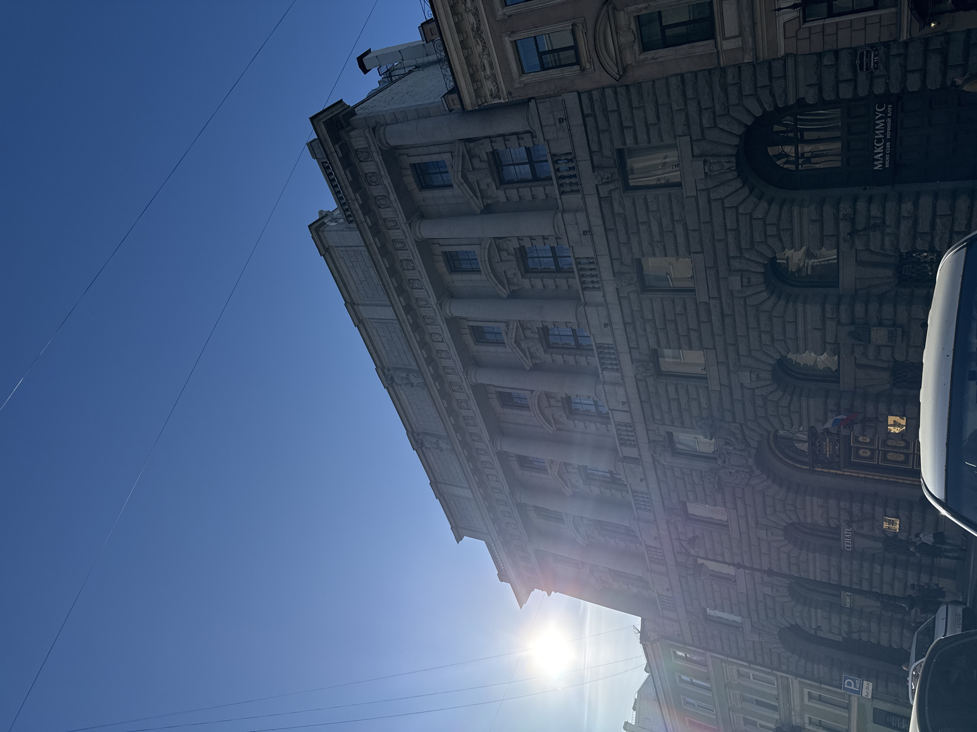 The image captures a low-angle, upward-looking view of a stately, multi-story building under a brilliant, clear blue sky in Saint Petersburg, Russia. The architecture suggests a historical or classical design, featuring rusticated stone block walls, prominent columns on the upper floors, ornate cornices, and a mix of rectangular and arched windows.
The sky is a uniform, deep blue, traversed by thin utility lines. The sun is directly visible, positioned high in the sky, causing a strong lens flare and illuminating the building's facade with bright light. This suggests clear weather and either midday or early afternoon.
Portions of several vehicles, including a white van or car, are visible along the right and bottom edges of the frame, indicating the building faces a street or parking area. No people are discernible.
Visible text on the building includes a sign for "МАКСИМУС" (MAKSIMUS) with "NIGHT CLUB НОЧНОЙ КЛУБ" beneath it, indicating a nightclub. Another sign, partially visible, reads "СЕНАТСКАЯ" (SENATSKAYA) and an address like "11-13" or "11-15," suggesting a street name and number. A larger vertical sign also shows "СЕНАТС" (SENATS). A small blue sign with a white "P" for parking is present. A Russian flag or national emblem is affixed to the building's exterior.