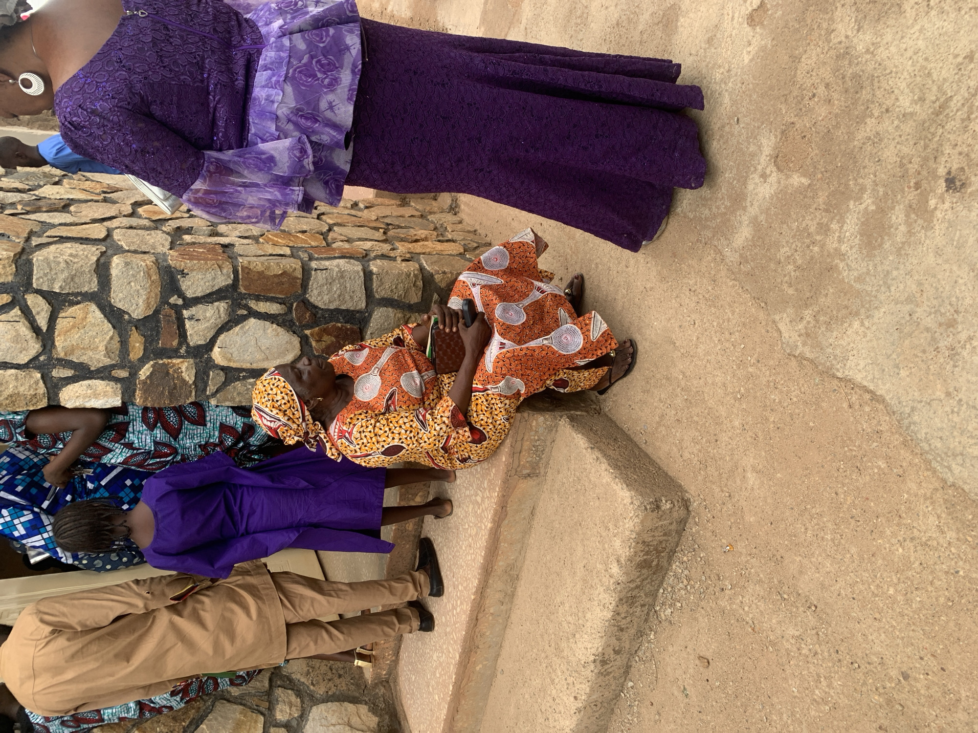 The image shows a group of people in what appears to be a public space in Jos, Nigeria.
There are two women sitting on concrete steps wearing brightly patterned African print dresses. One is clad in orange and yellow, and the other is seated behind her on the steps.
Another woman stands nearby wearing a long purple lace dress with a lighter purple sash. To the left are two individuals: one dressed in a purple top and the other in a tan outfit. The surface is primarily concrete, with stone walls also visible.