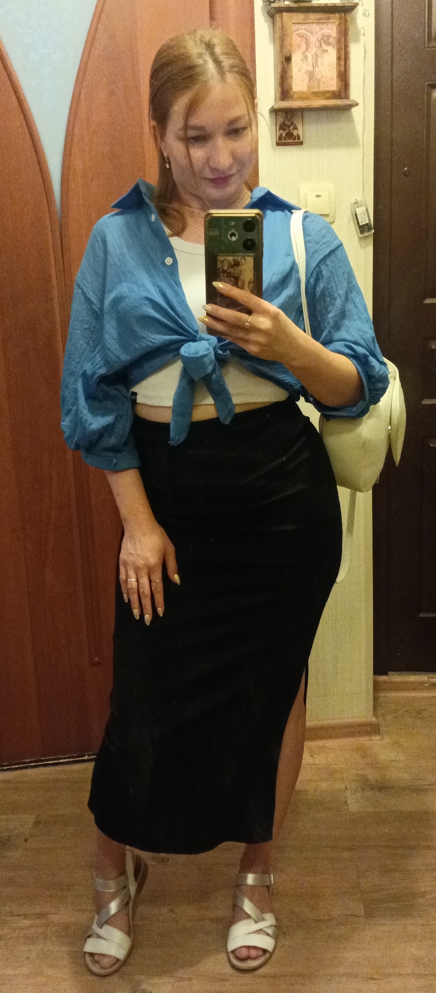 A woman is posing for a selfie in front of a mirror in Unknown, Unknown. She is wearing a white crop top, a blue button-down shirt tied at the waist, a black midi skirt with a slit, and white sandals with silver straps. She is holding her phone in her right hand and has a white backpack slung over her left shoulder. The background includes a brown door, a light-colored wall with a small decorative shelf and a light switch, and a dark brown door. The floor is light brown.